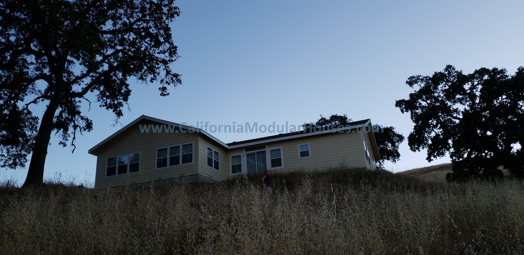 A house on a hillside with dry grass and trees, under a clear sky, partially silhouetted against the light.  Bay Area Prefab Homes, Modular Homes in California.