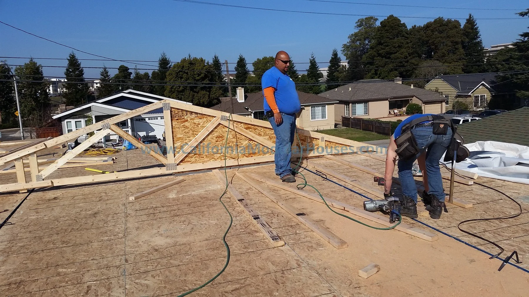 Two construction workers building a roof on a house, with one standing and the other bending over, surrounded by construction tools and wood framing. Bay Area Prefab Modular Home, Modular Home CA,