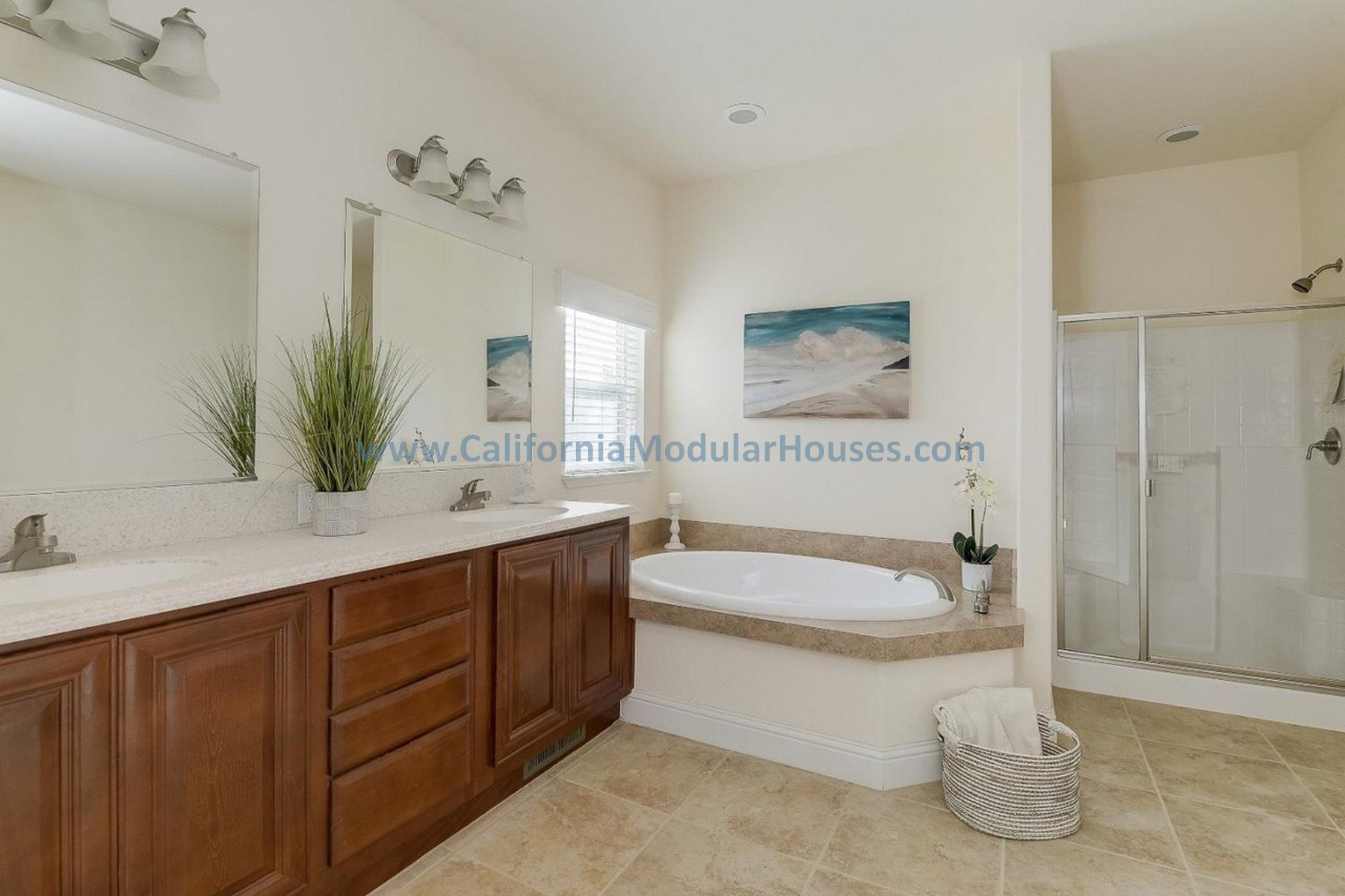 Bathroom with dual sinks, large mirror, potted plant, bathtub with beige surround, framed beach artwork, window with blinds, basket with towels, shower stall with glass door, beige tiled flooring. Modular Home CA,