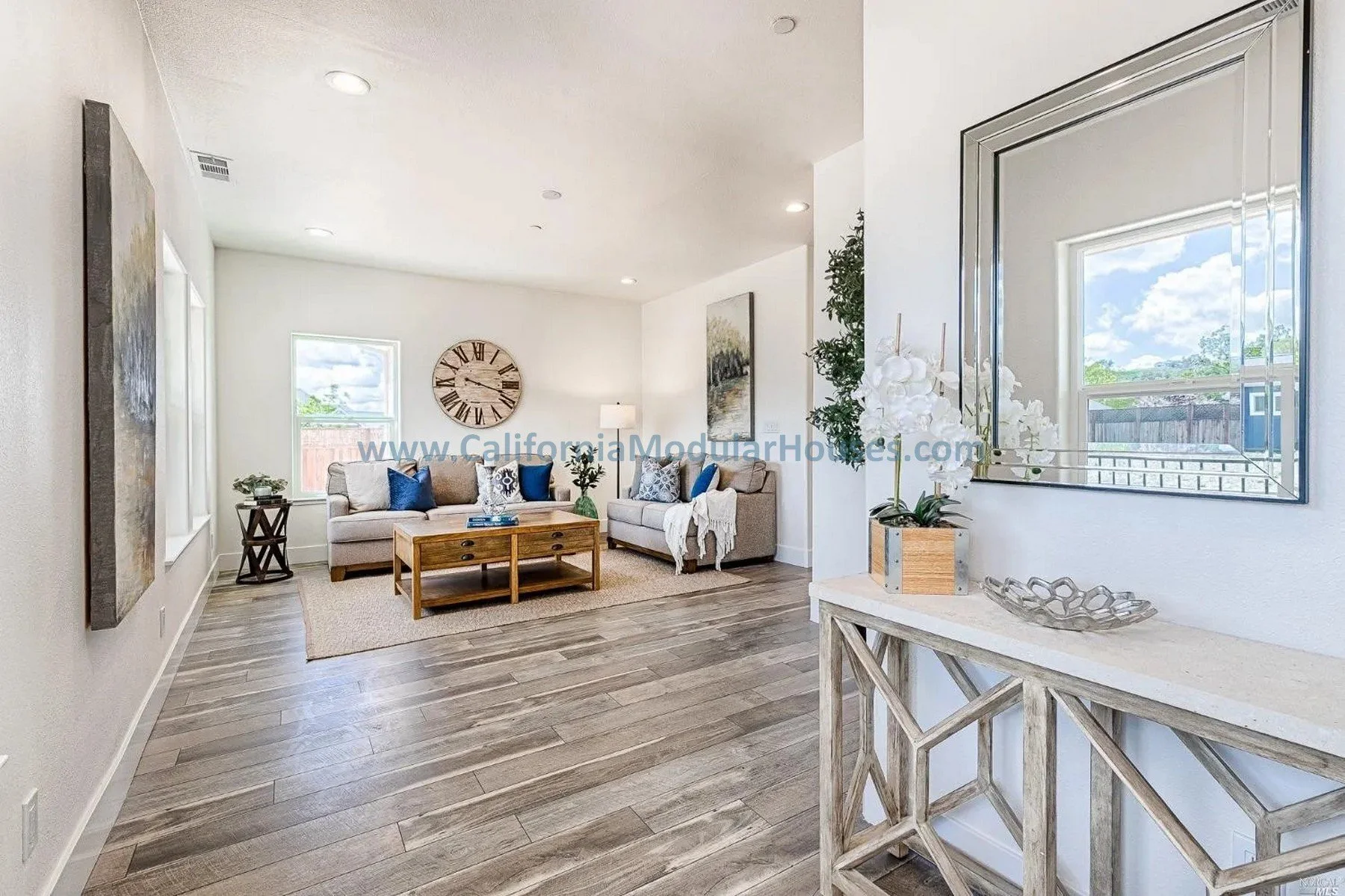 Living room with two beige couches, a wooden coffee table, a large wall clock, artwork, and decorative pillows, with natural light coming from windows.