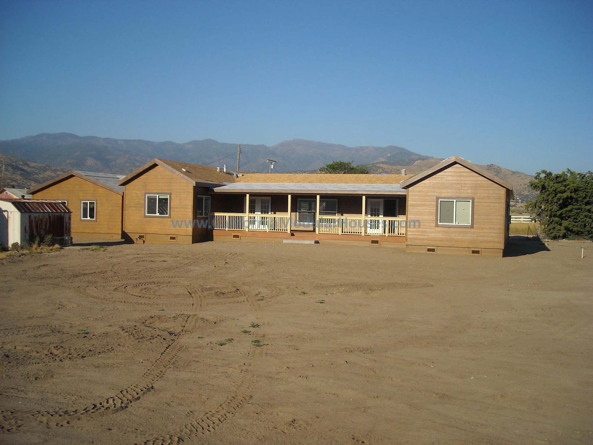 Newly constructed single-story house with a wooden exterior, large windows, and a small front porch set against a background of mountains and clear blue sky. Remaining ground in front of the house is bare dirt.