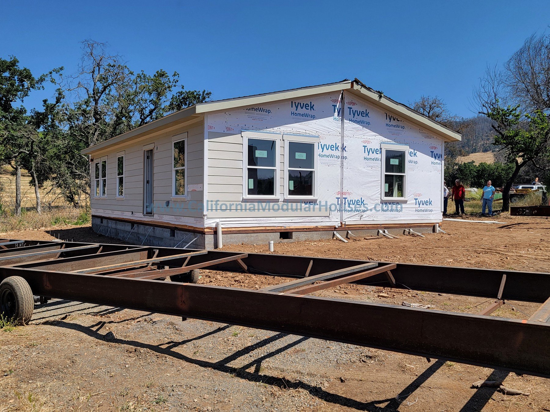 A house under construction with white siding and multiple windows, sitting on a foundation with no landscaping around. There is a trailer in the foreground and a few workers and trees in the background.