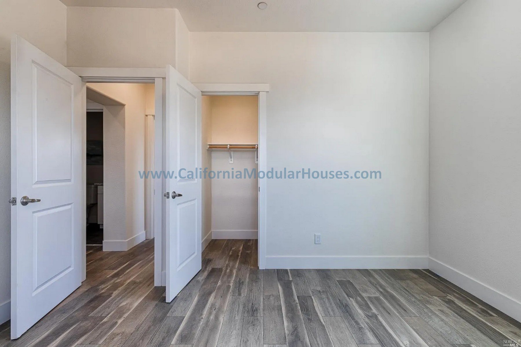 Empty bedroom with white walls, hardwood flooring, closet with wooden rod, open door leading to bathroom