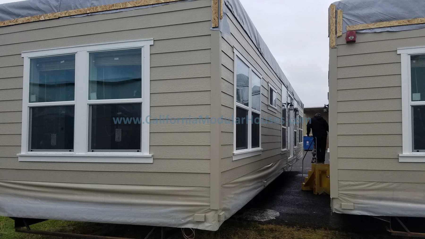 Two beige prefabricated houses with white-framed windows are being assembled in an outdoor setting.   Modular Home CA, California Modular Homes, Prefab Modular Homes California, California Modular, California Modular Home.