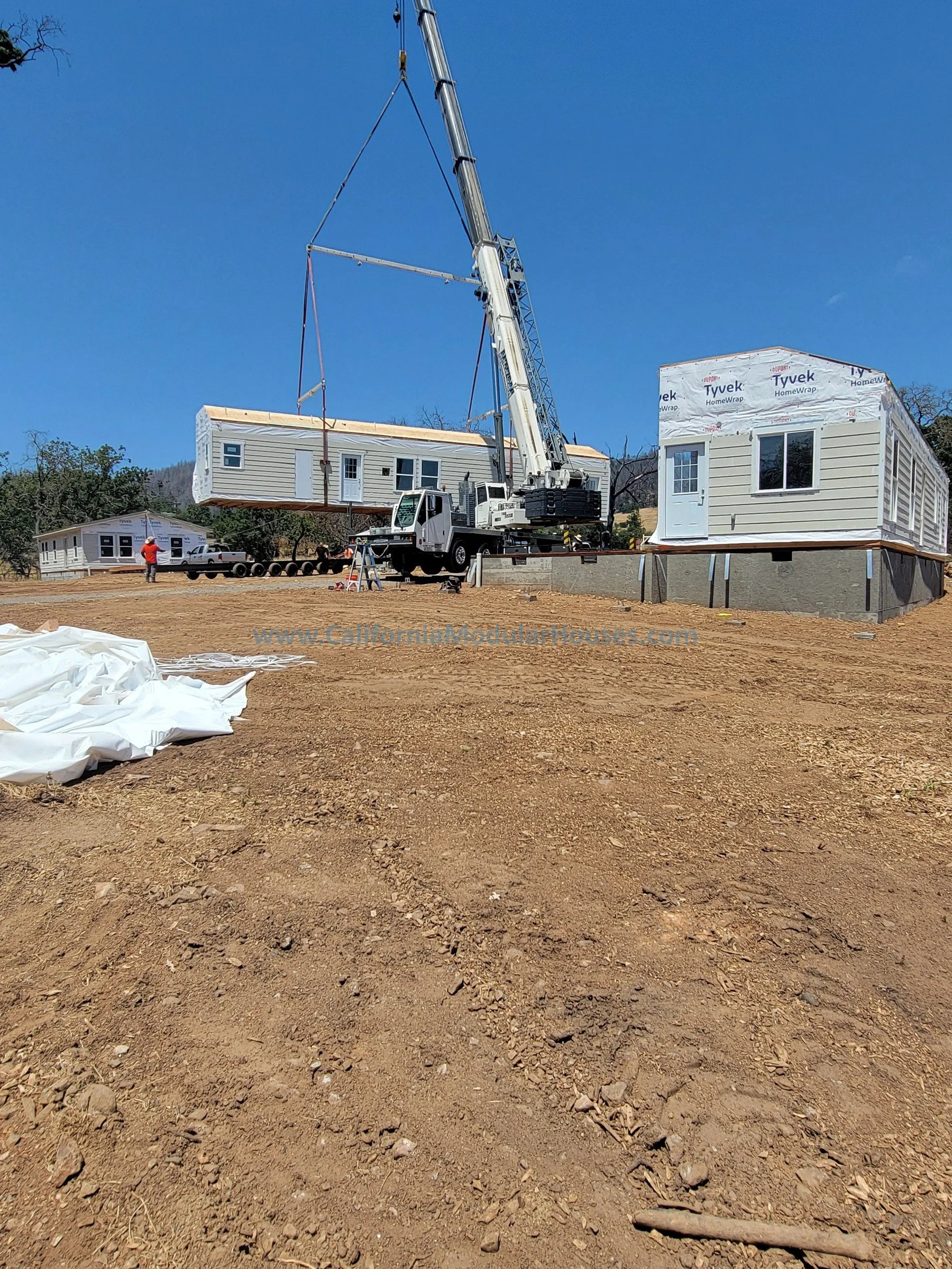 Construction site where a crane is lifting a prefabricated modular home into place, with partially assembled modular homes built on a dirt lot under a clear blue sky.