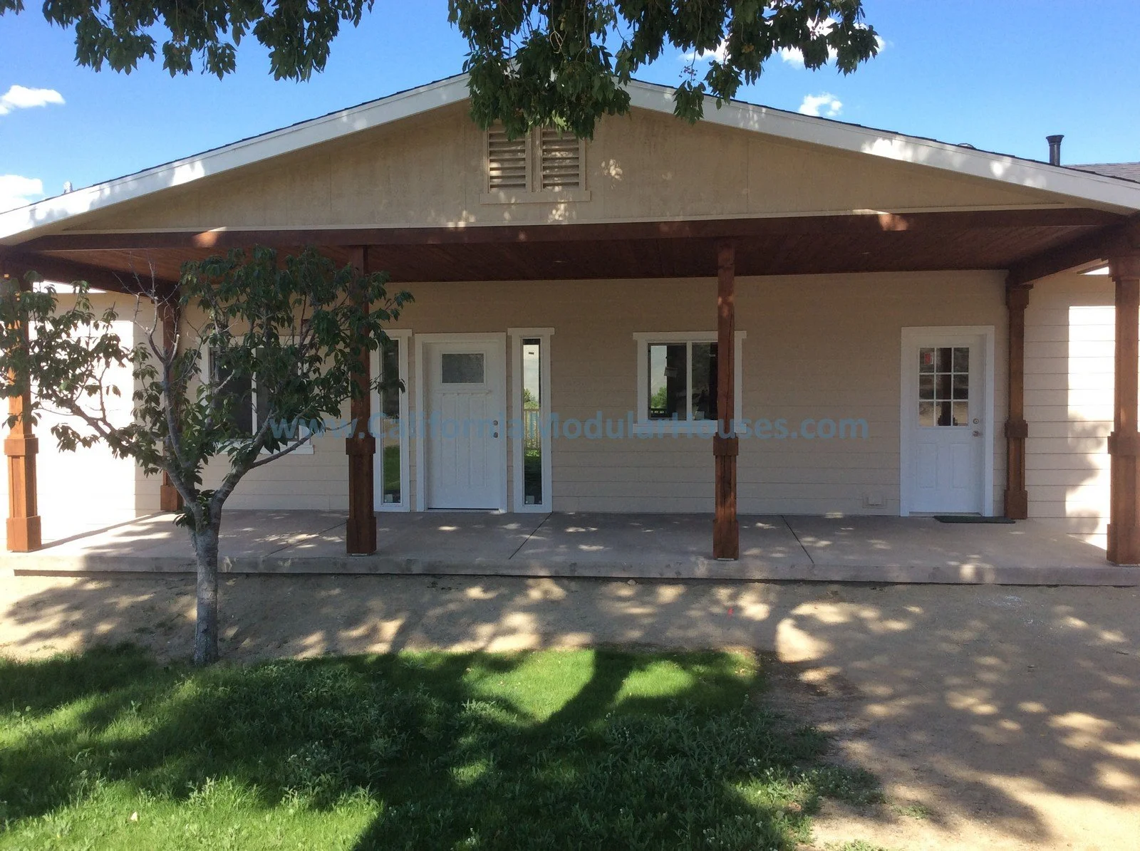 Front view of a house with a covered porch, two doors, a small window, a tree, green lawn, and shadow patterns on the ground under a partly cloudy sky.