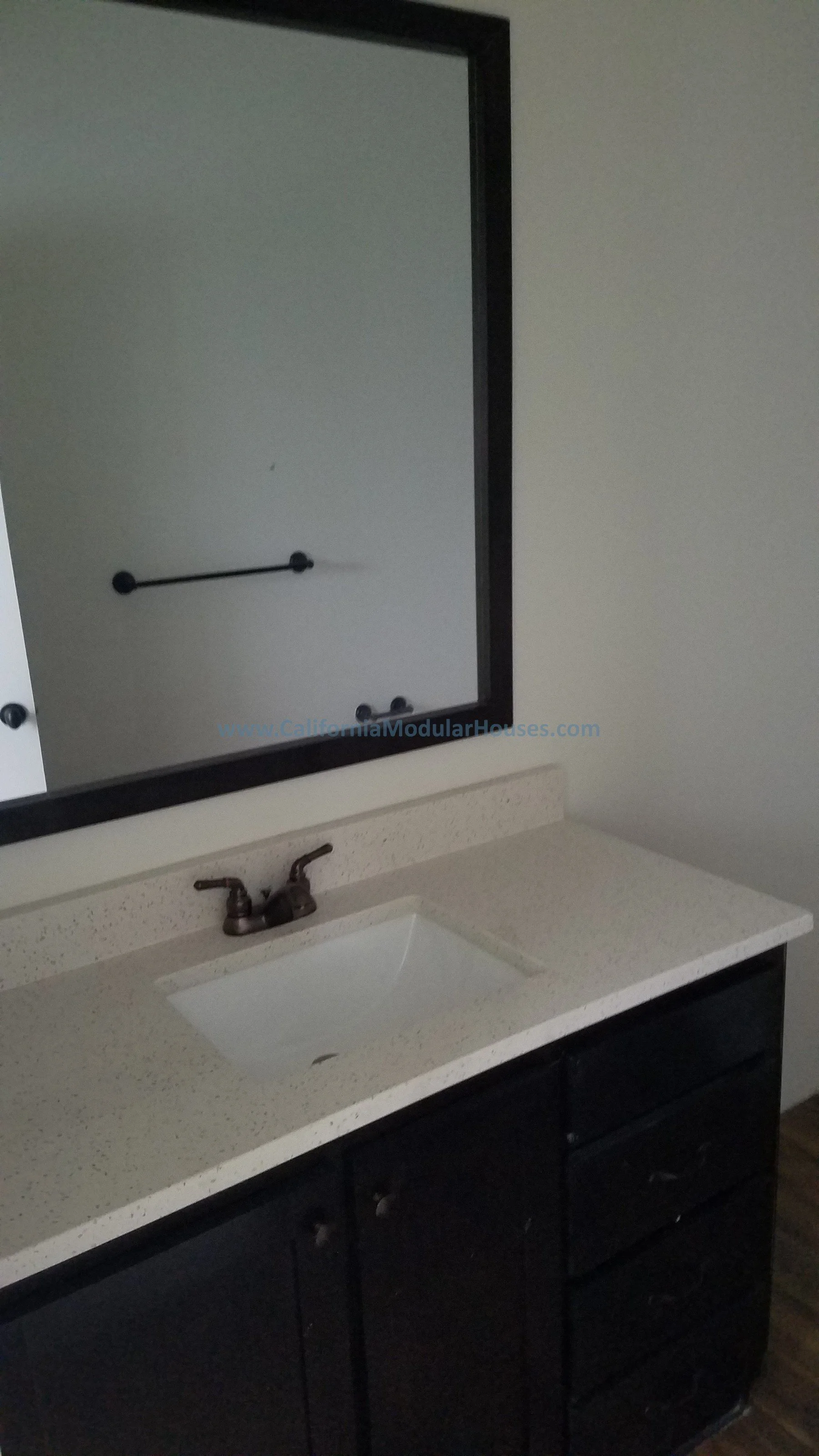 Bathroom vanity with a speckled countertop, a white sink, bronze faucet, black cabinet, and a large mirror with a black frame.  Pre-Fabricated Homes, Prefab Home California, Modular Homes in California,