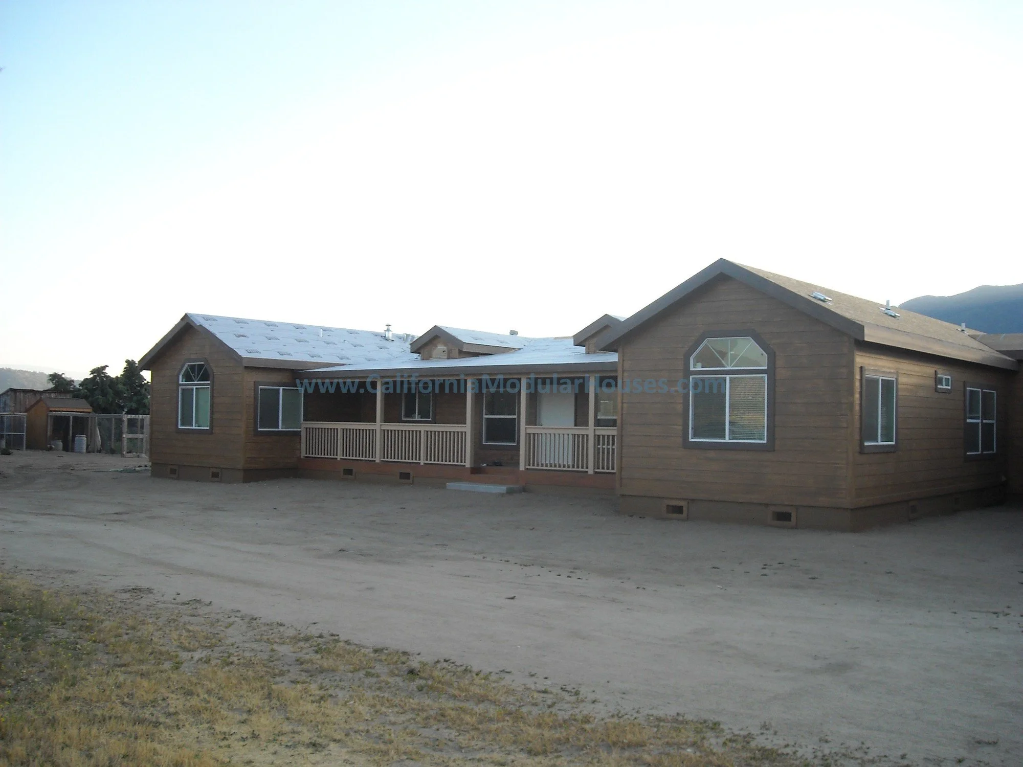A newly constructed single-story wooden house with multiple gabled roofs in a rural area, with dirt ground in front and a small shed to the left.