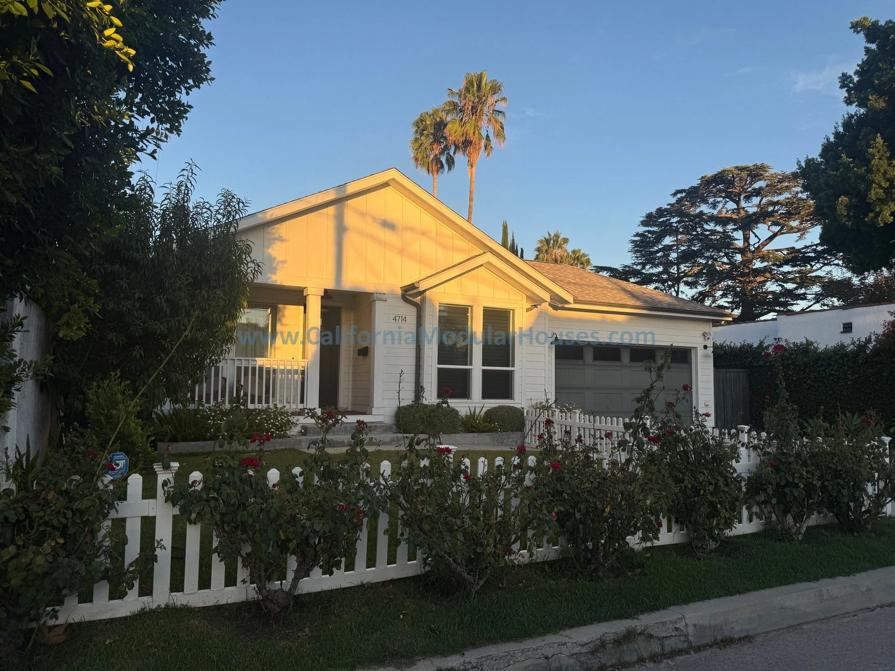A white house with a lawn and white picket fence, surrounded by trees and bushes, with the sun setting and casting a warm glow.
