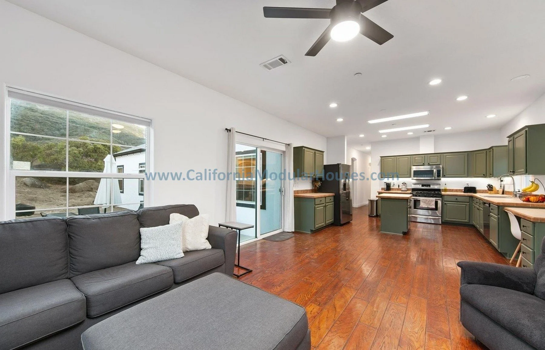 Living room with gray sofa and pillows, open kitchen with green cabinets, stainless steel appliances, hardwood floors, large window, sliding glass door, ceiling fan, and recessed lighting.
