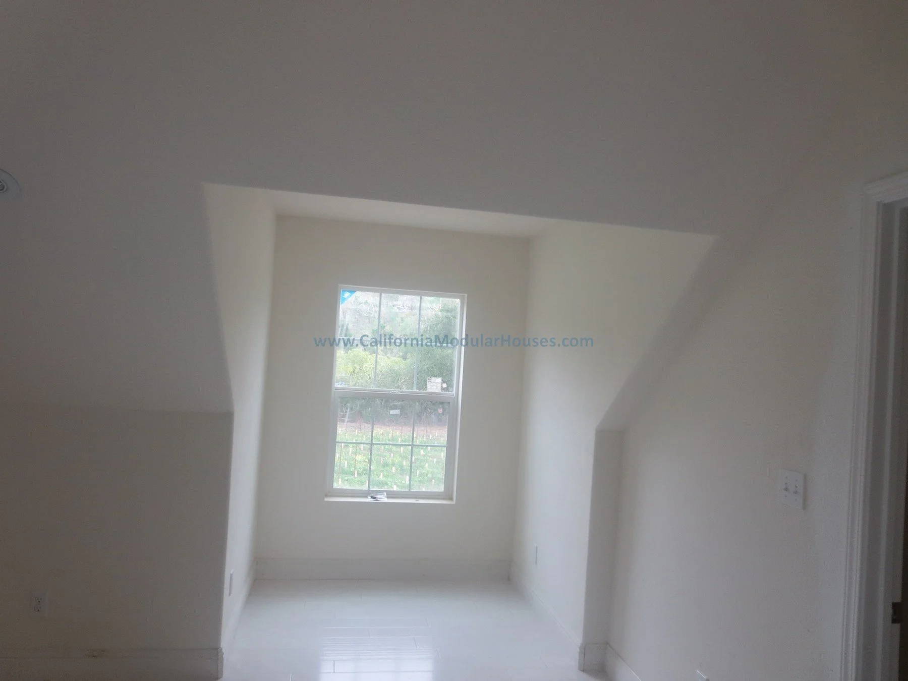 Empty room with a window showing outdoor greenery and a cloudy sky, white walls, and ceiling with sloped architectural features.  Napa County, CA Vineyard.  Modular homes.  SFR.  New House Cape Cod.  Unfinished attic.  