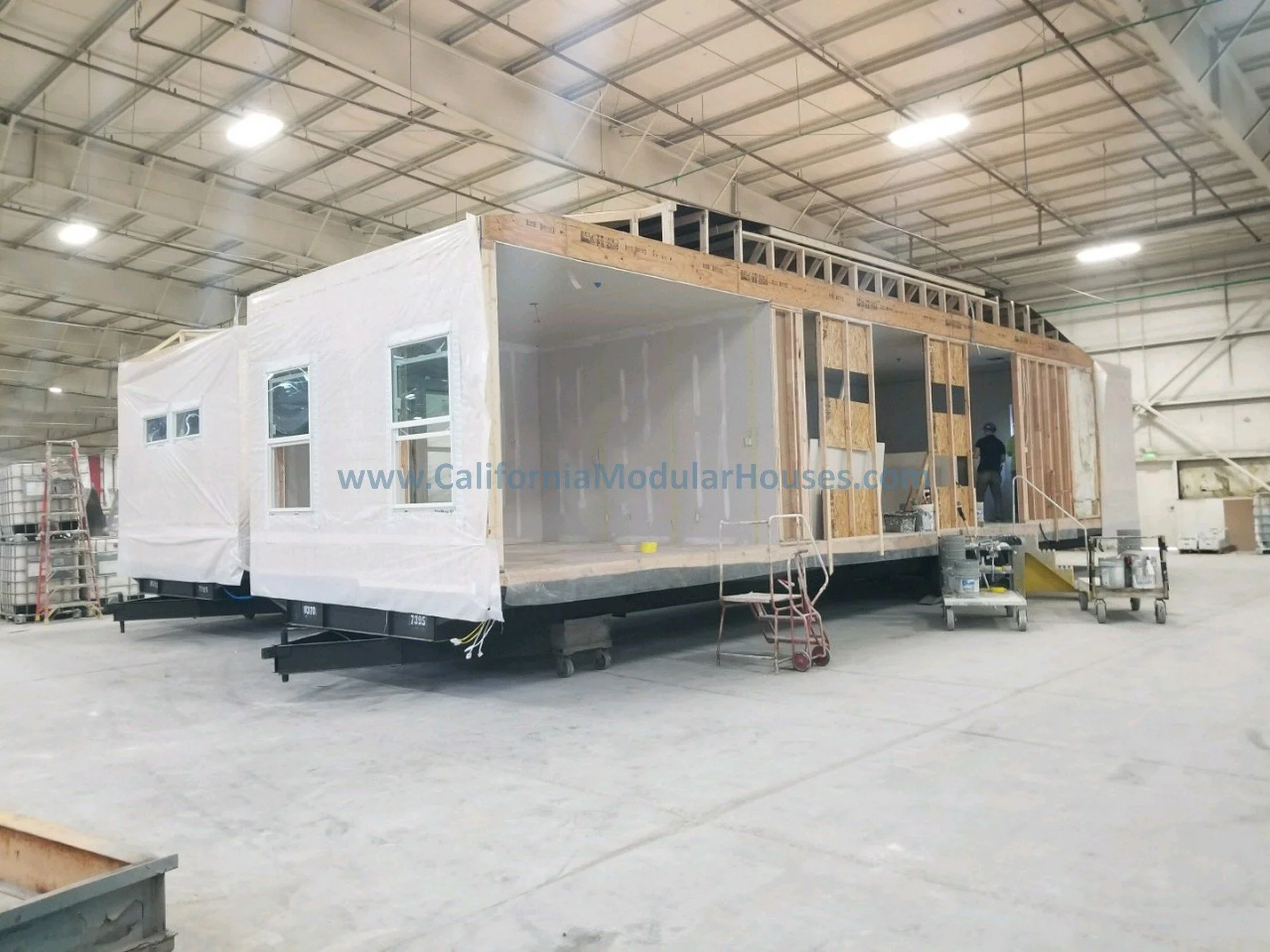 Interior of a modular house under construction inside a factory, with exposed wooden framing, window openings, and construction tools and materials nearby.  Modular Home CA, California Modular Homes, Prefab Modular Homes California,