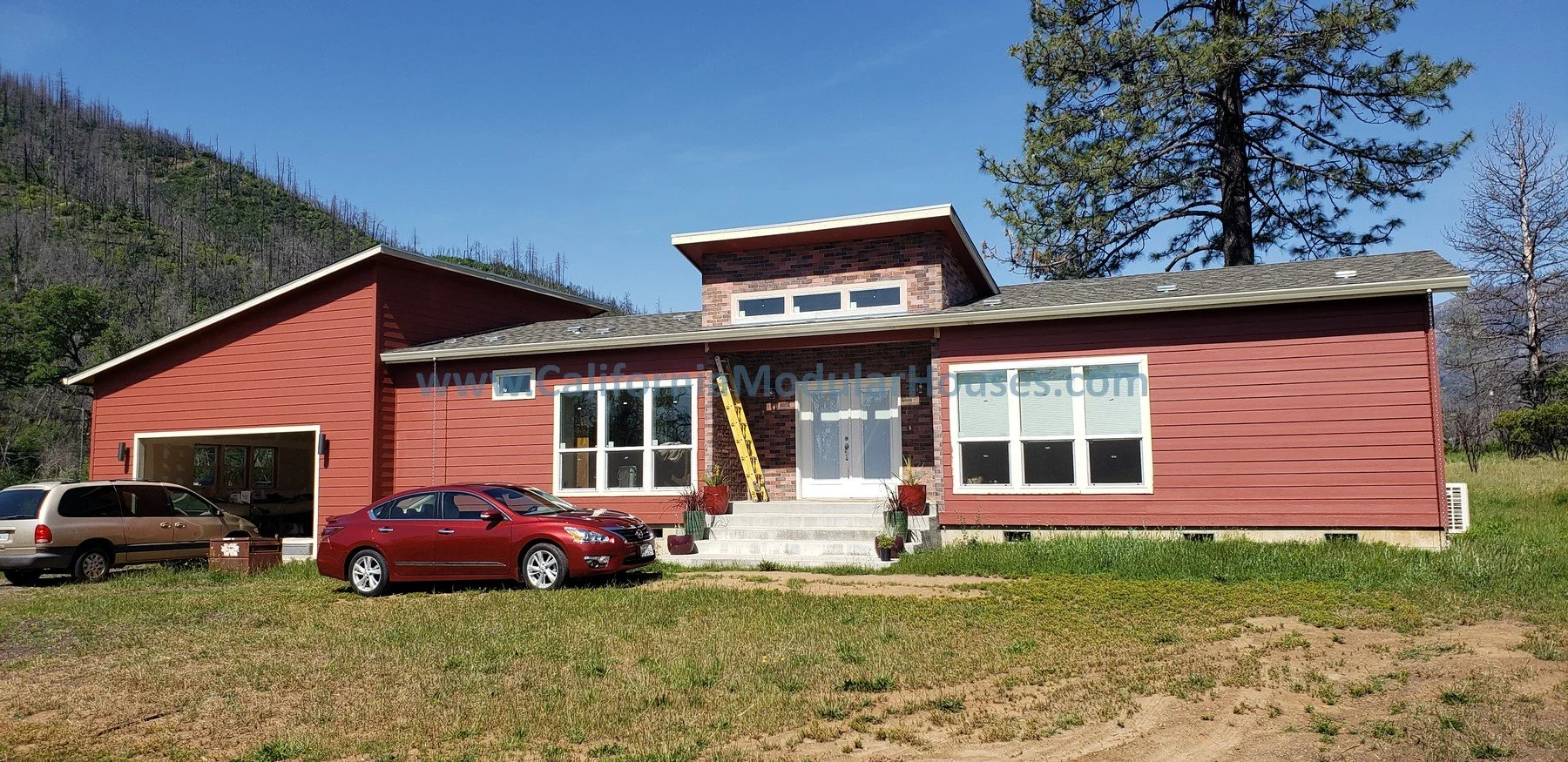 Modern house with red siding, brick chimney, and multiple large windows, surrounded by a grassy yard with cars parked in front, set against a hillside with trees and a clear blue sky. Middletown Prefab Homes, Middletown, Lake County, CA.
