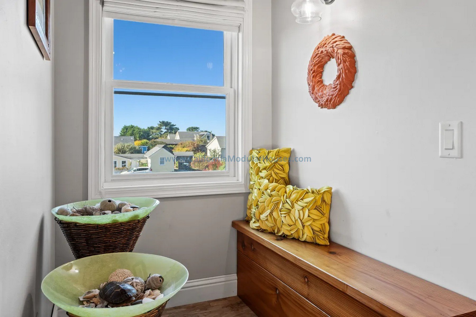 A small window with a view of neighborhood houses and blue sky, white walls with a pink wreath, two yellow pillows with textured patterns on a wooden bench, two baskets filled with shells and decorative objects, and a light switch.  Modular home CA.