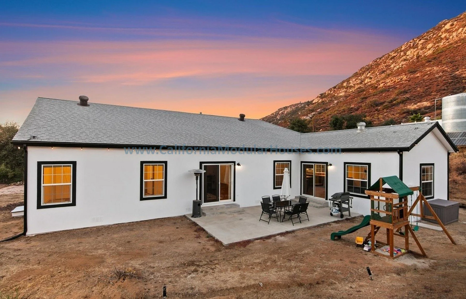 Modern white house with black window frames and a gray shingled roof, outdoor patio with table, chairs, umbrella, and grill, a small playset with slide and sandbox, mountainous landscape in the background at sunset.