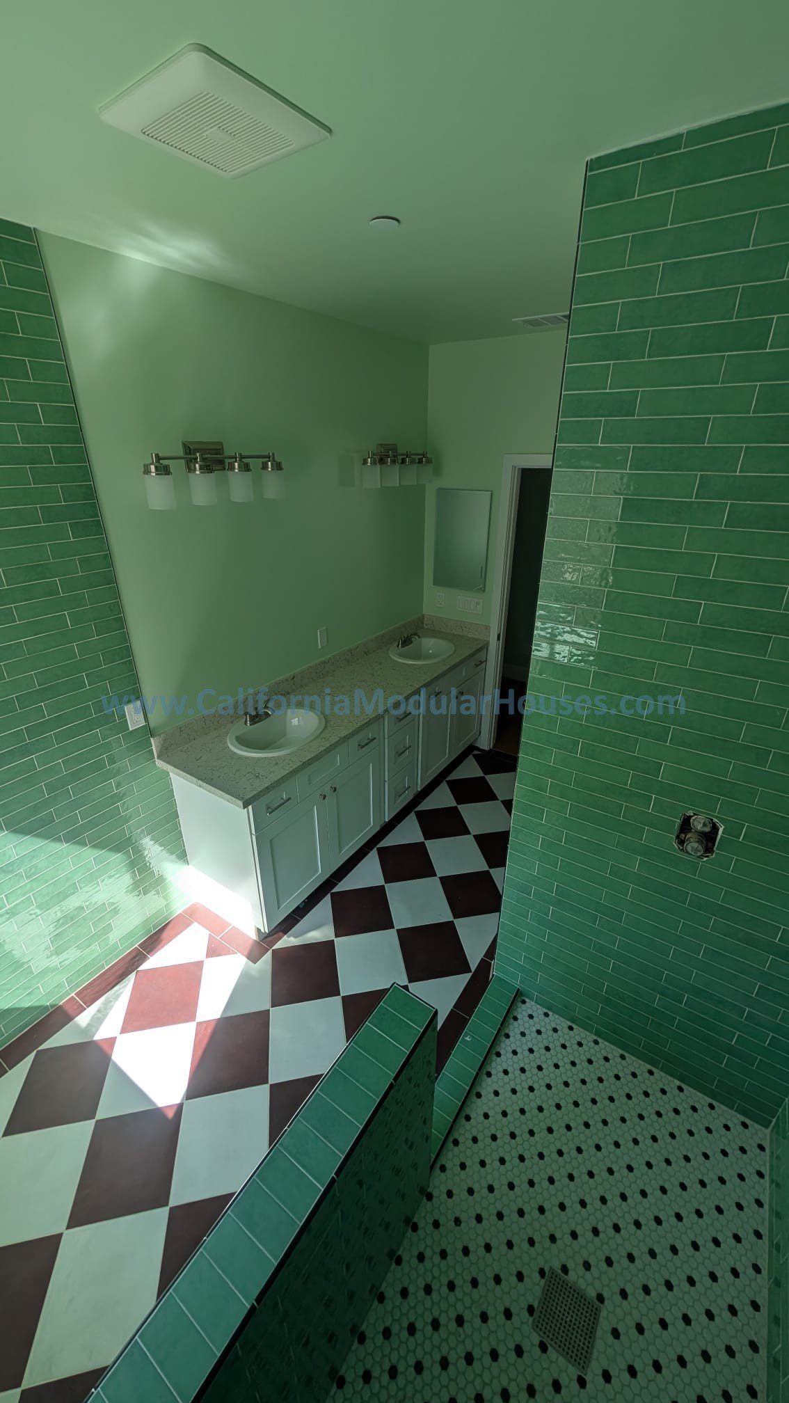 View of a bathroom with green tiled walls, a twin sink vanity with a granite countertop, black and white checkered floor, and a separate walk-in shower area with black and white small hexagon tiles.