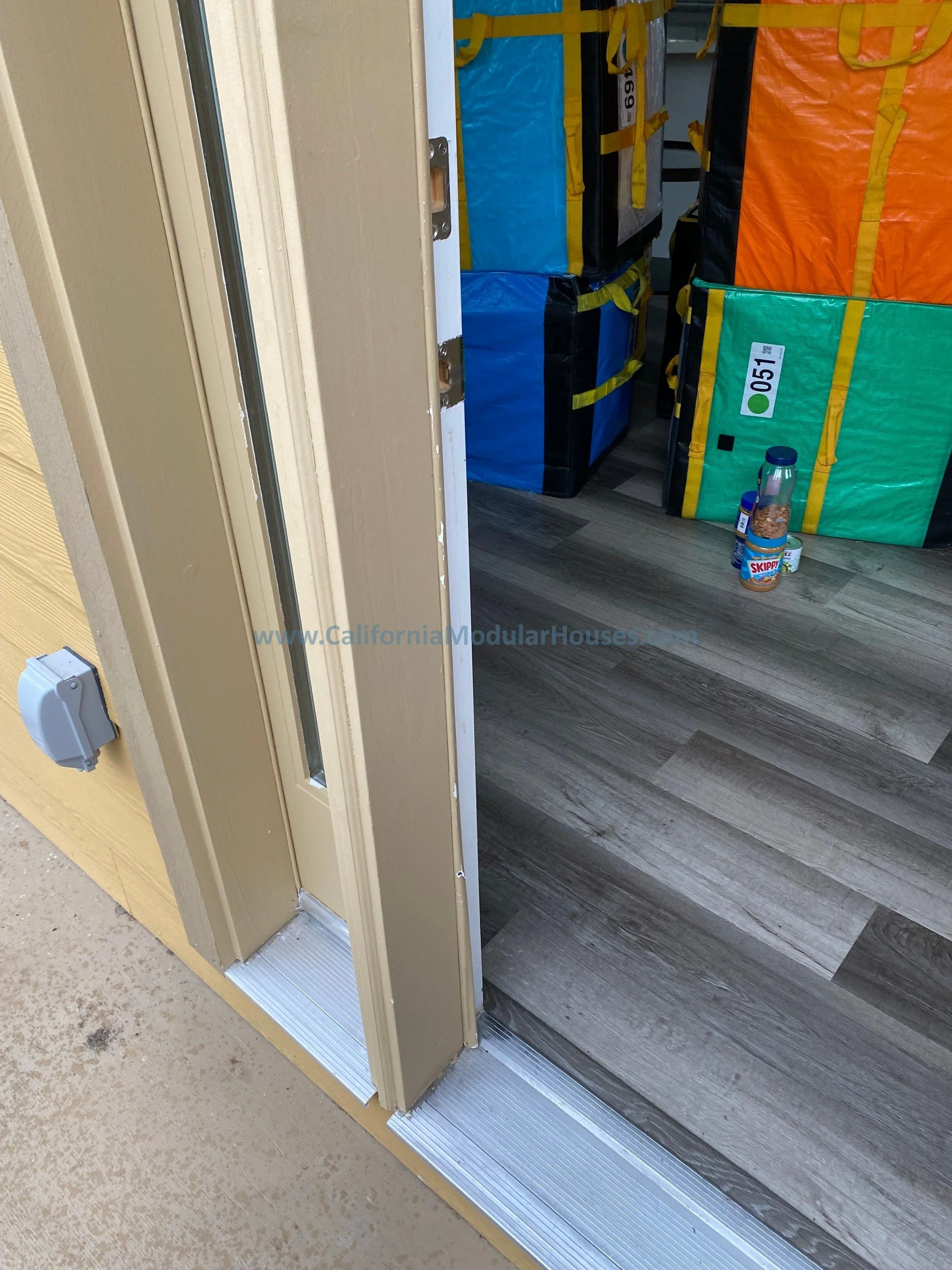 View of an open door leading to a room with folded colorful fabric storage bags, a bottle of iced coffee, a small container, and a jar of snacks on the floor. Modular Accessory Dwelling Unit  Prefab.