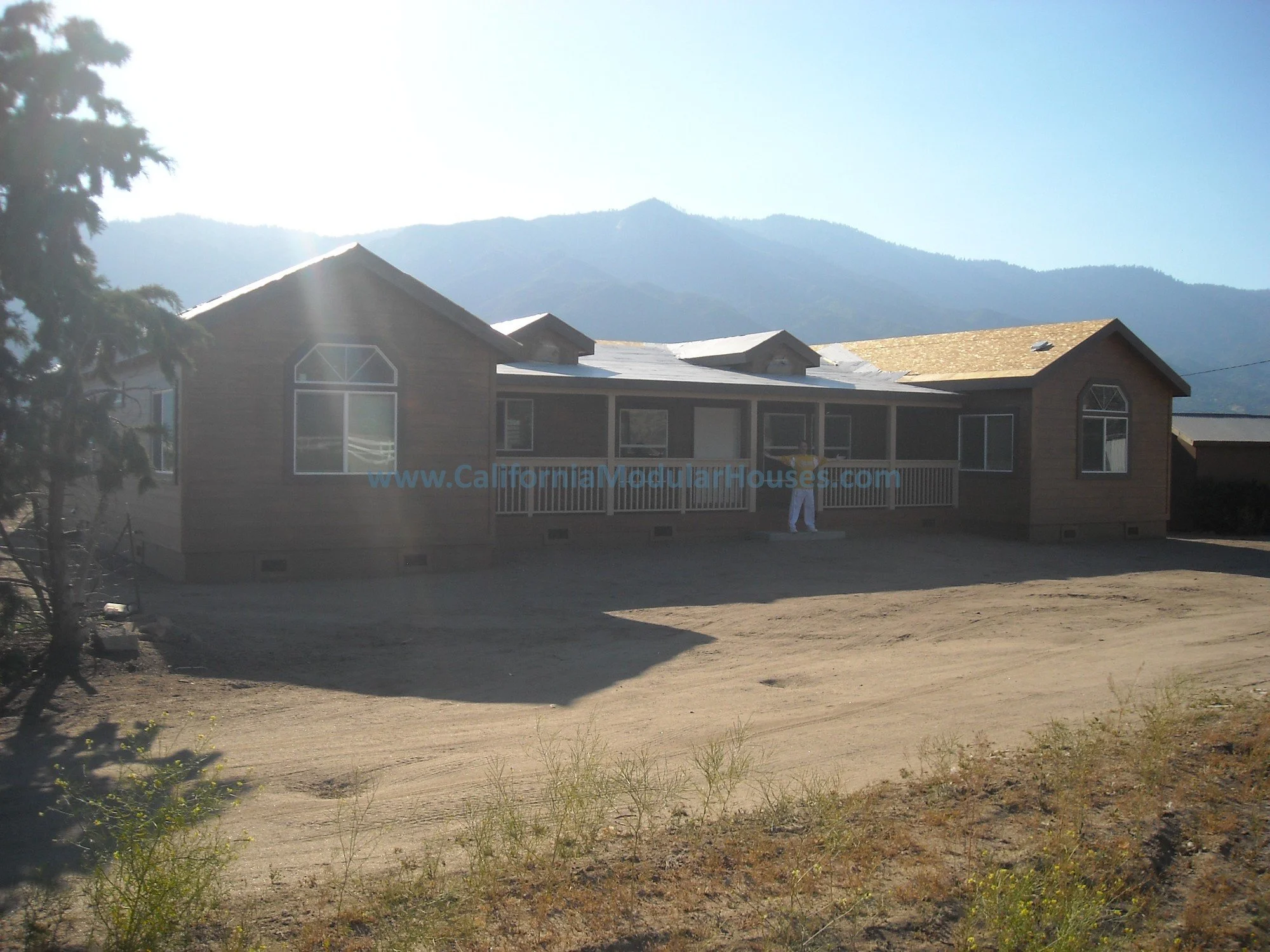 A single-story prefab house under completion with a wooden exterior appearance, a front porch, large windows, and a mountainous landscape in the background.  