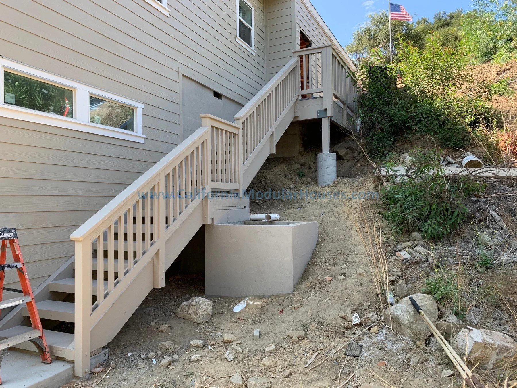 Construction of an outdoor staircase with beige railings leading up a slope next to a beige house with horizontal siding and small windows.  Modular Home in California, Modular Home California, Modular Homes.