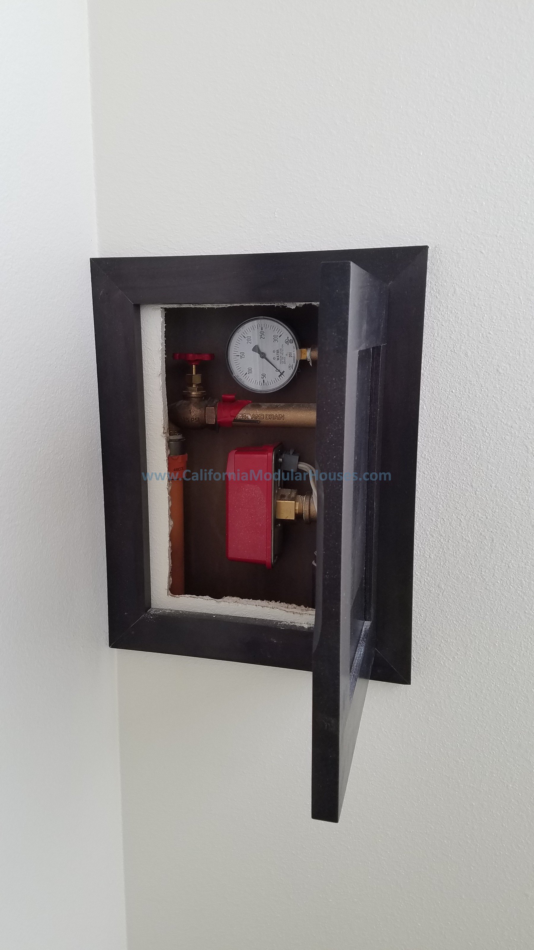 View of an access panel with plumbing and a pressure gauge installed on a white wall for mandatory fire sprinklers.  Modular Home CA, California Modular Homes, Prefab Modular Homes California,