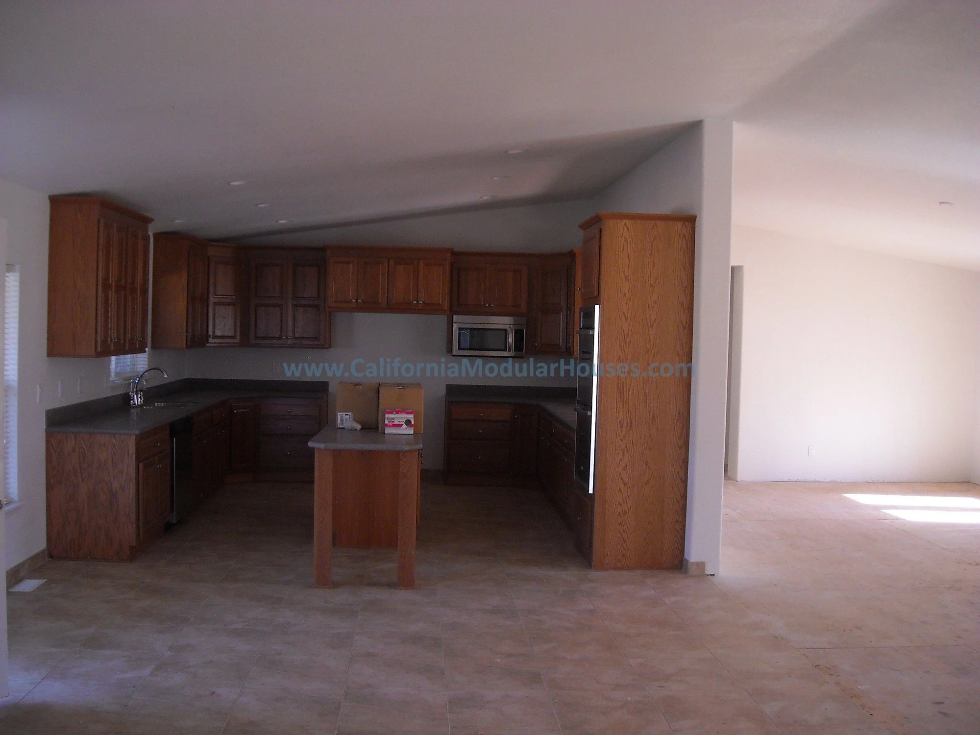 Empty kitchen with wooden cabinets, gray countertops, and a small central island. Adjacent room has light-colored walls and sunlight streaming in.  Modular Home, Kern County, California.  