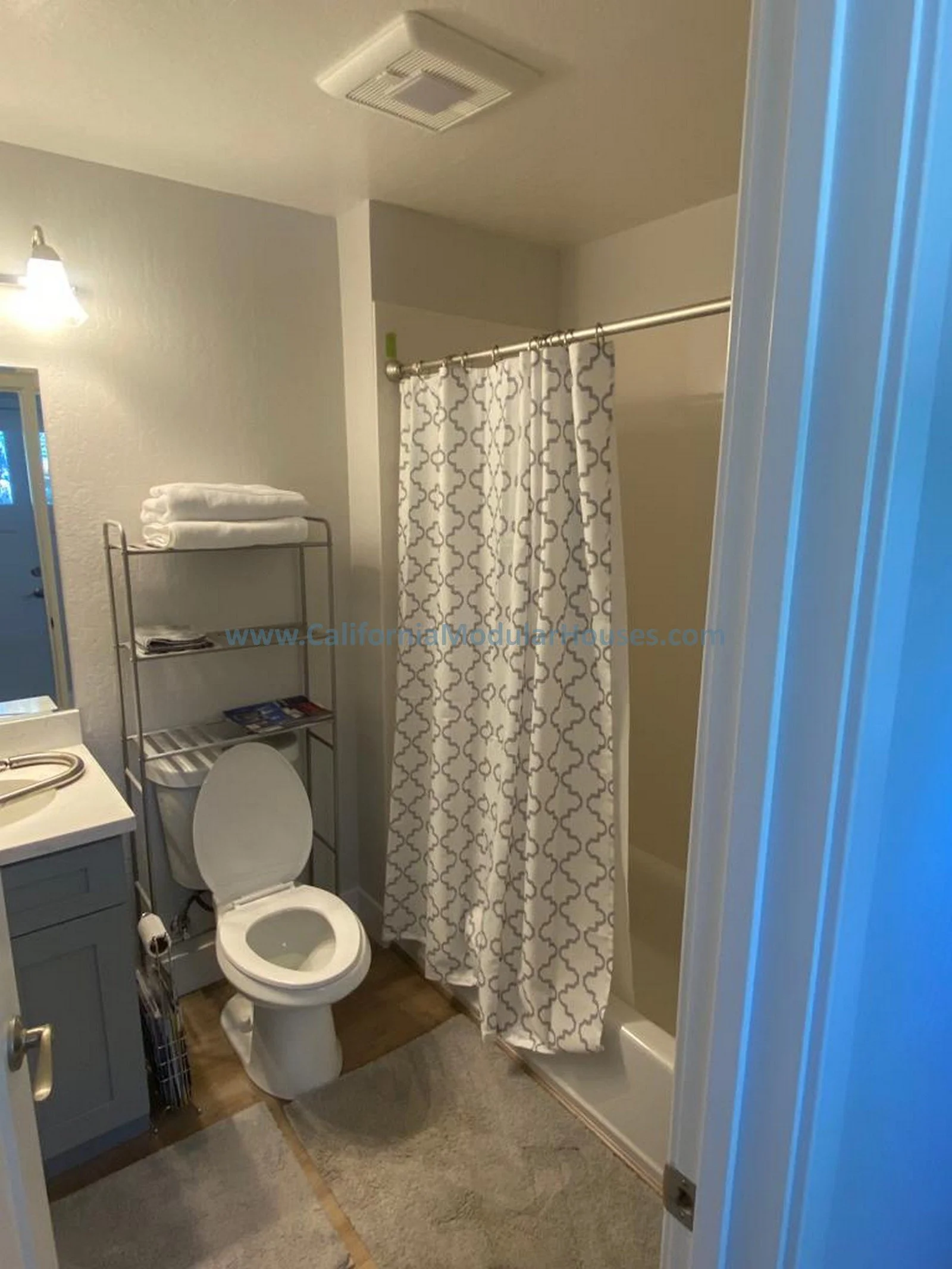 Small bathroom with a toilet, a sink, a metal shelf with towels, and a shower with a patterned curtain.