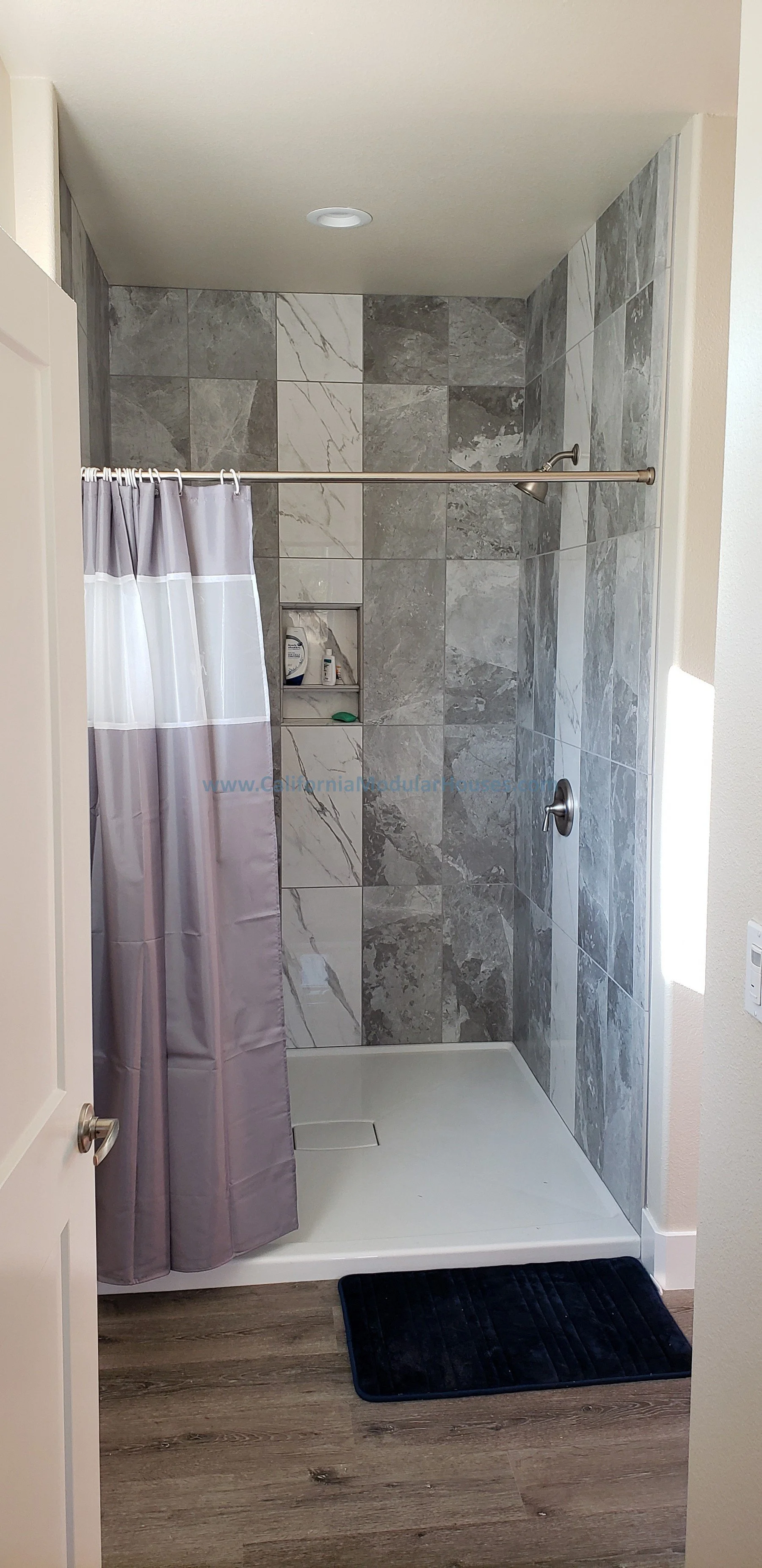 A modern walk-in shower with gray marble tiles, a rain shower head, a small built-in shelf with toiletries, a white shower base, a purple and white shower curtain, and a black bath mat on wood flooring.  Prefab Modular ADU