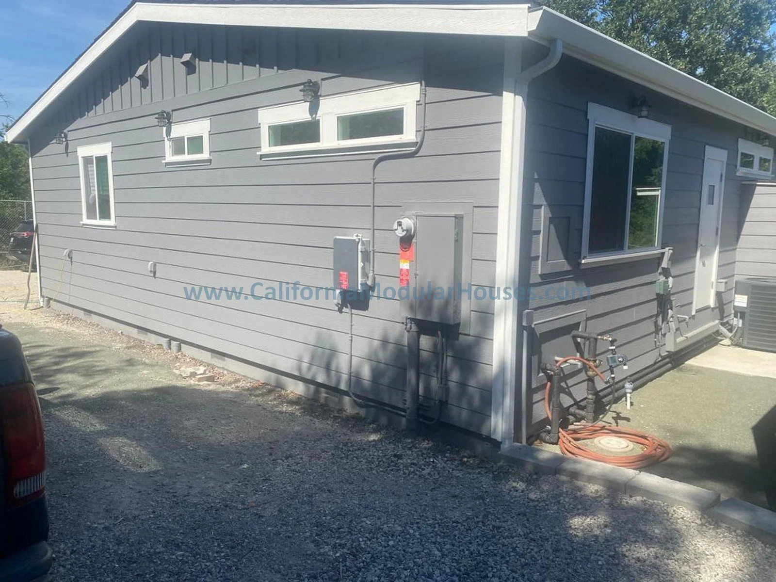 Gray modular house with exterior electrical and utility connections, windows, and a gravel parking lot.