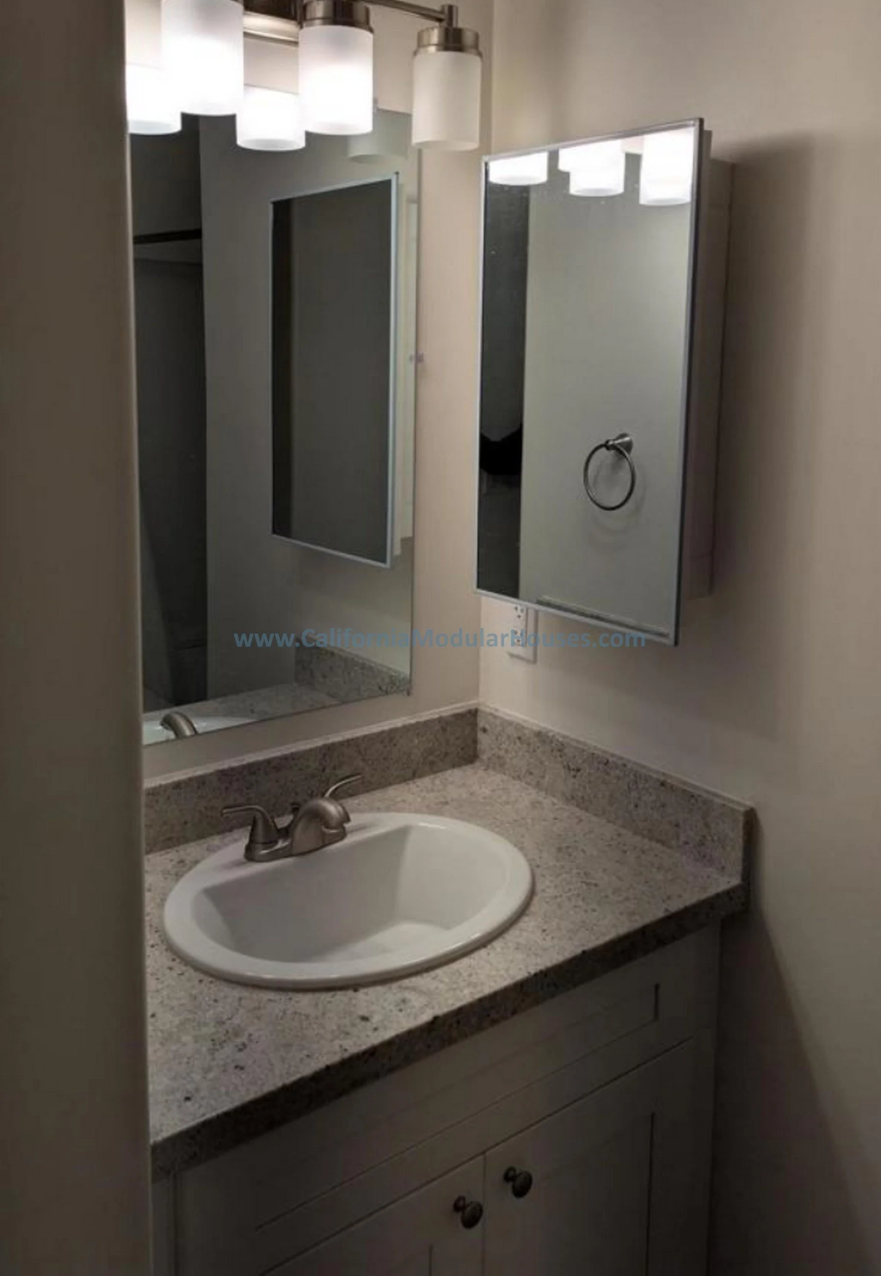 Bathroom with a granite countertop, a white oval sink, a wall mirror, a light fixture with three cylindrical shades, a towel ring, and cabinetry below the countertop.  Prefab Modular ADU, Modular Home CA