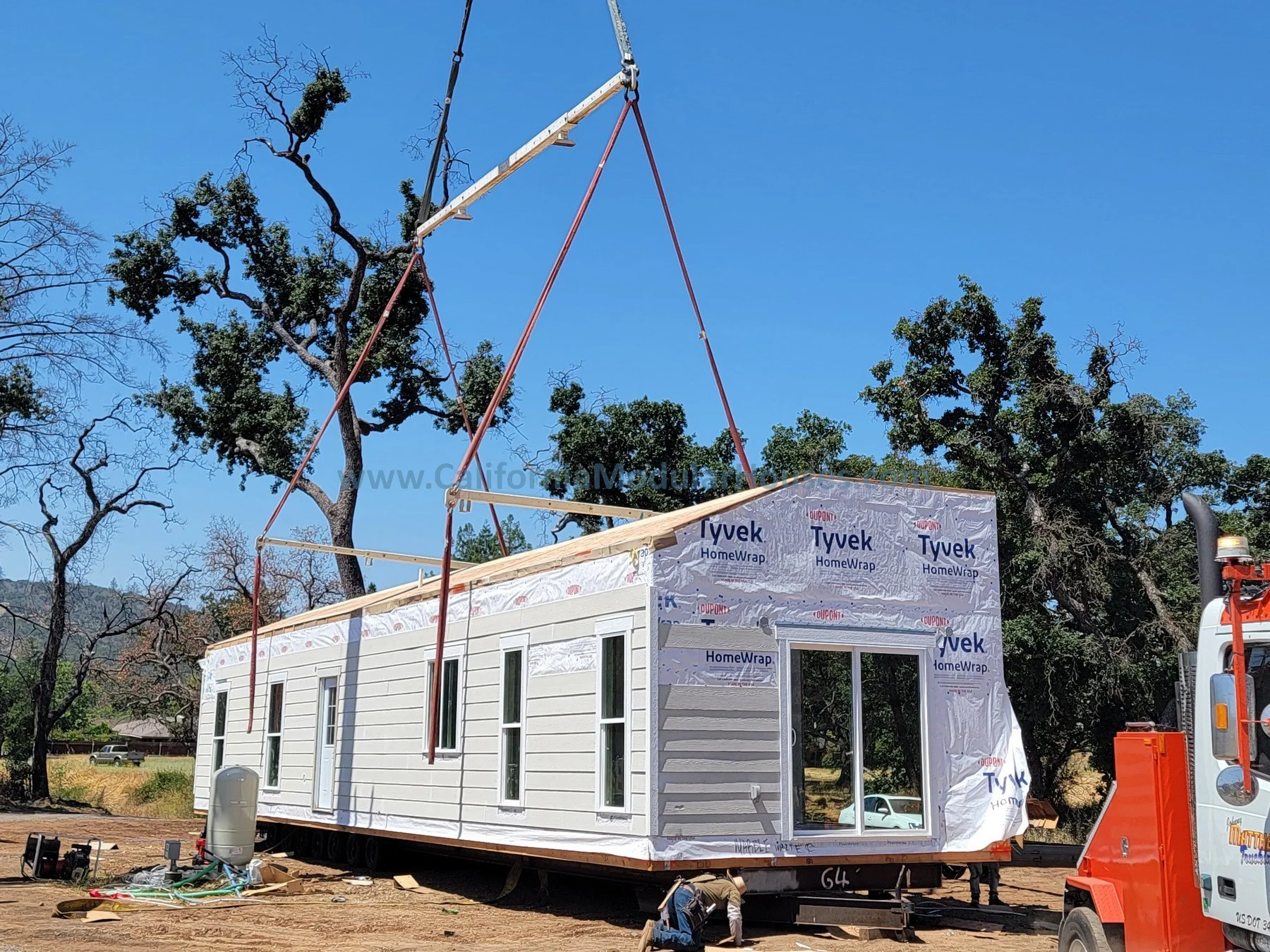 A tiny house under construction with Tyvek HomeWrap on the exterior, placed on a trailer. A crane is lifting roof trusses into position, and there is construction equipment nearby on the ground.