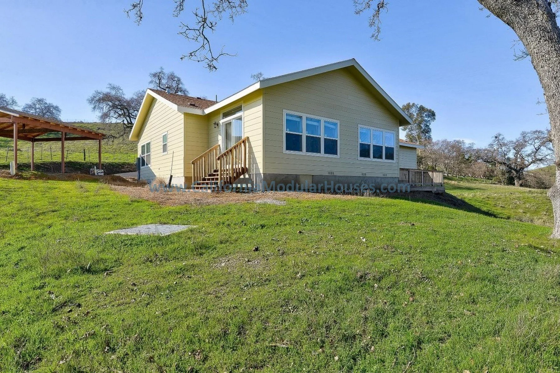 A yellow modular house with a small wooden staircase, situated on a grassy hillside with a few large trees and a clear blue sky. Bay Area Prefab Modular Home, Modular Home CA, California Modular Homes,