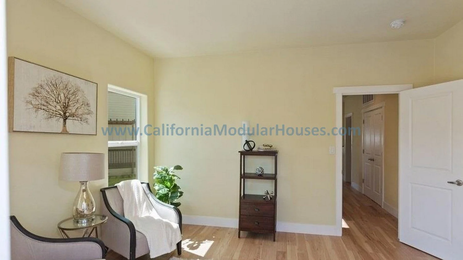 California modular home.  prefab.  A living room with beige walls, a window, a framed tree print, a table lamp, two armchairs, a small shelving unit, and a doorway leading to a hallway.