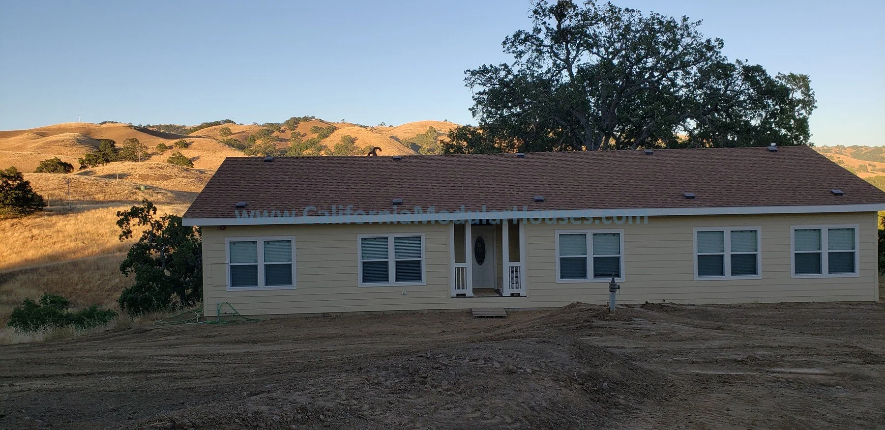 Newly constructed, yellow single-story house with a brown roof, multiple windows, a small front porch, and a surrounding dirt yard, situated in a hilly landscape with trees and dry grass hills in the background. Modular Home CA,