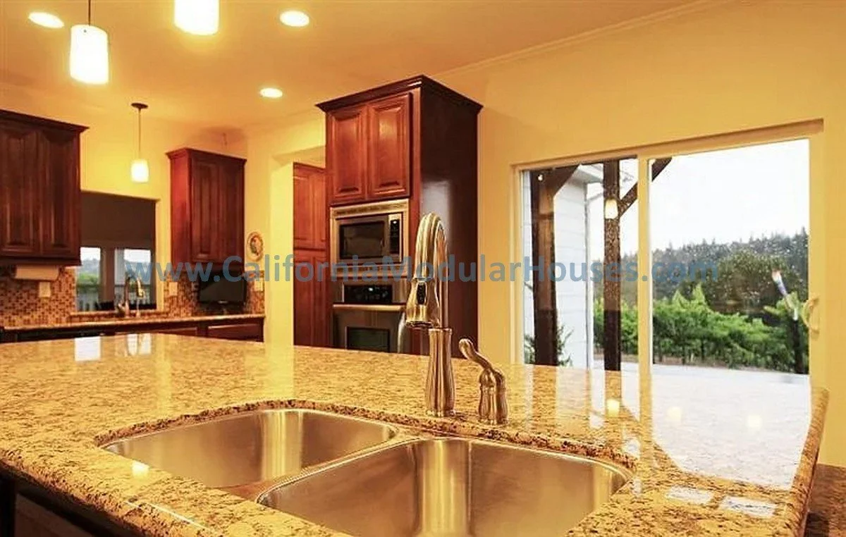 Modern kitchen with granite island counter, dual sinks, wooden cabinets, stainless steel faucet, microwave, oven, and slide door leading to an outdoor area with greenery.