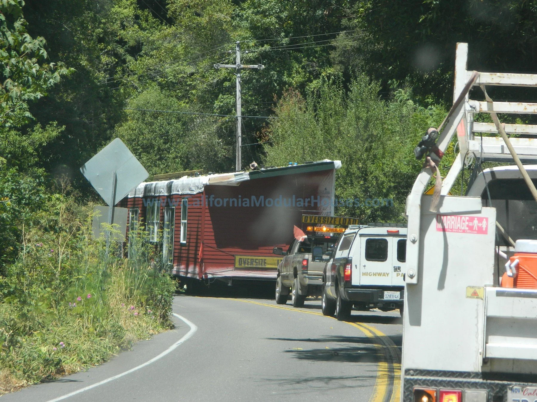 A large red semi-truck with an oversized load moving along a winding road, surrounded by green trees, with multiple vehicles following behind and utility poles visible in the background. Lake County Modular, Northern California Prefab Homes.