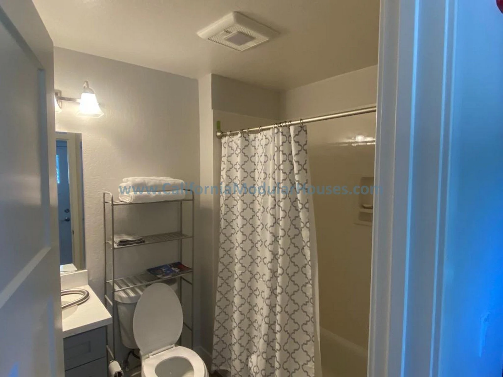 Small bathroom with a toilet, a metal shelf with folded towels, a shower with a patterned shower curtain, a small mirror, a sink, and a wall-mounted light fixture.