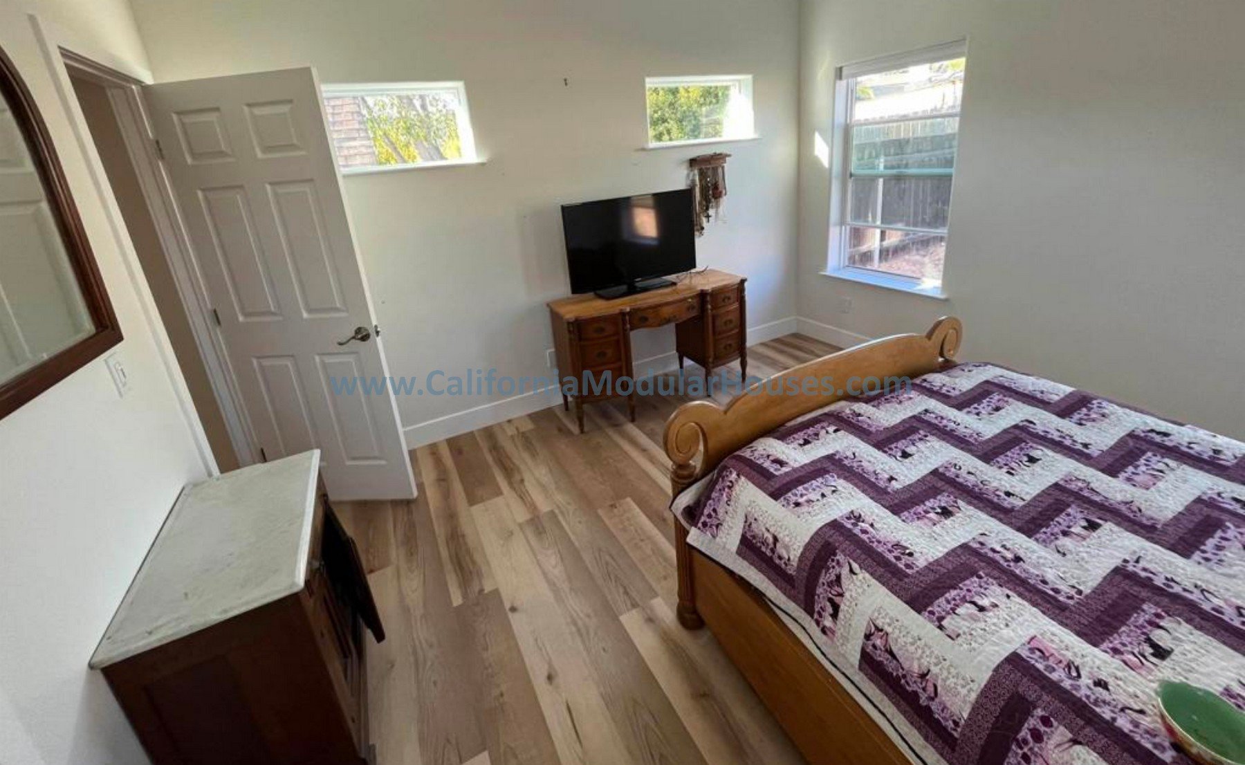 Three windows, a small wooden dresser with a flat-screen TV on top, a wall hanging, and a white door.  San Diego County, CA. Prefab Modular ADU, Oceanside CA.  Accessory Dwelling Unit.  California Modulars. 