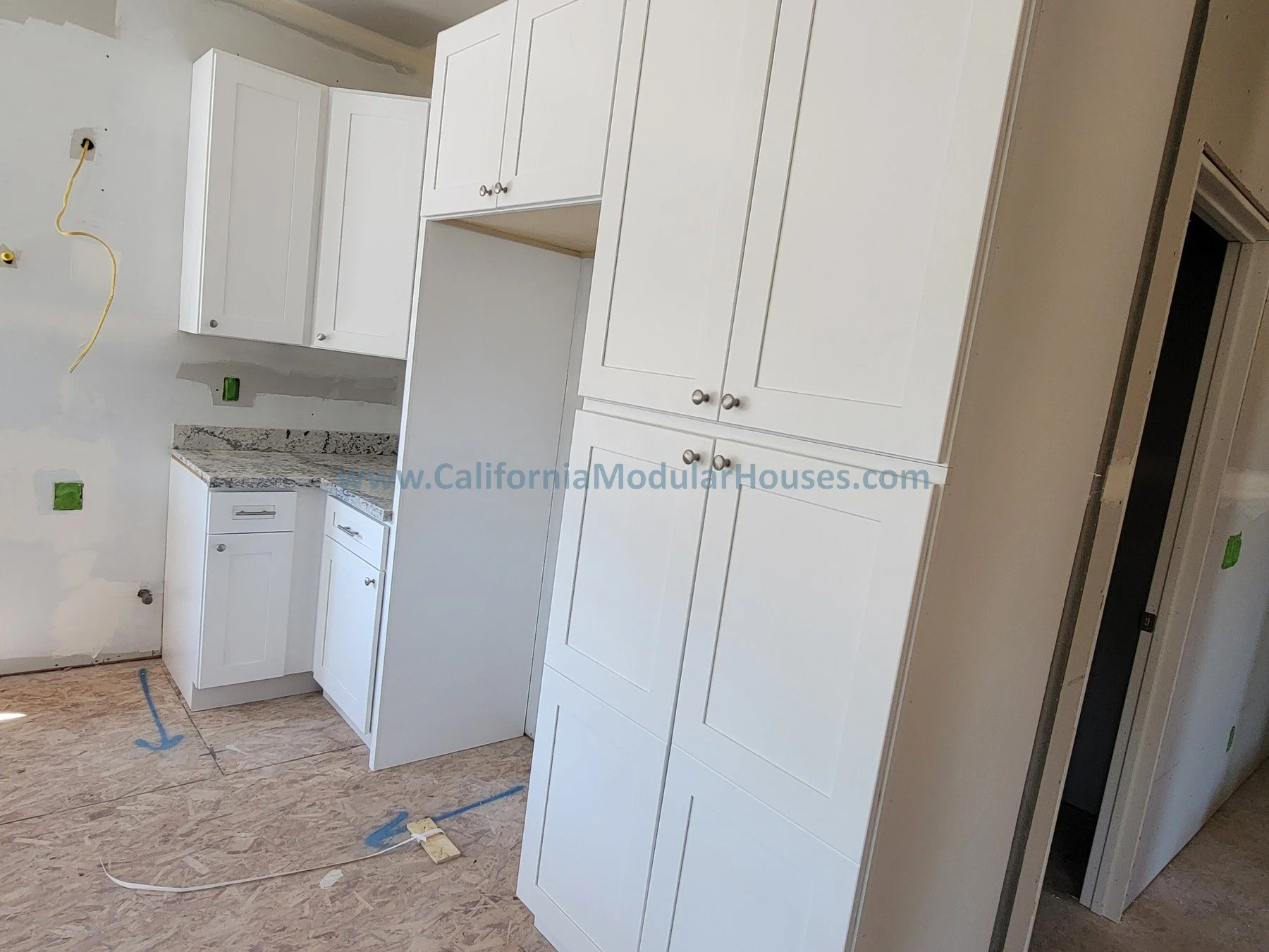 Kitchen with white cabinets and granite countertops under construction, with uninstalled appliances, electrical wires, and unfinished walls.