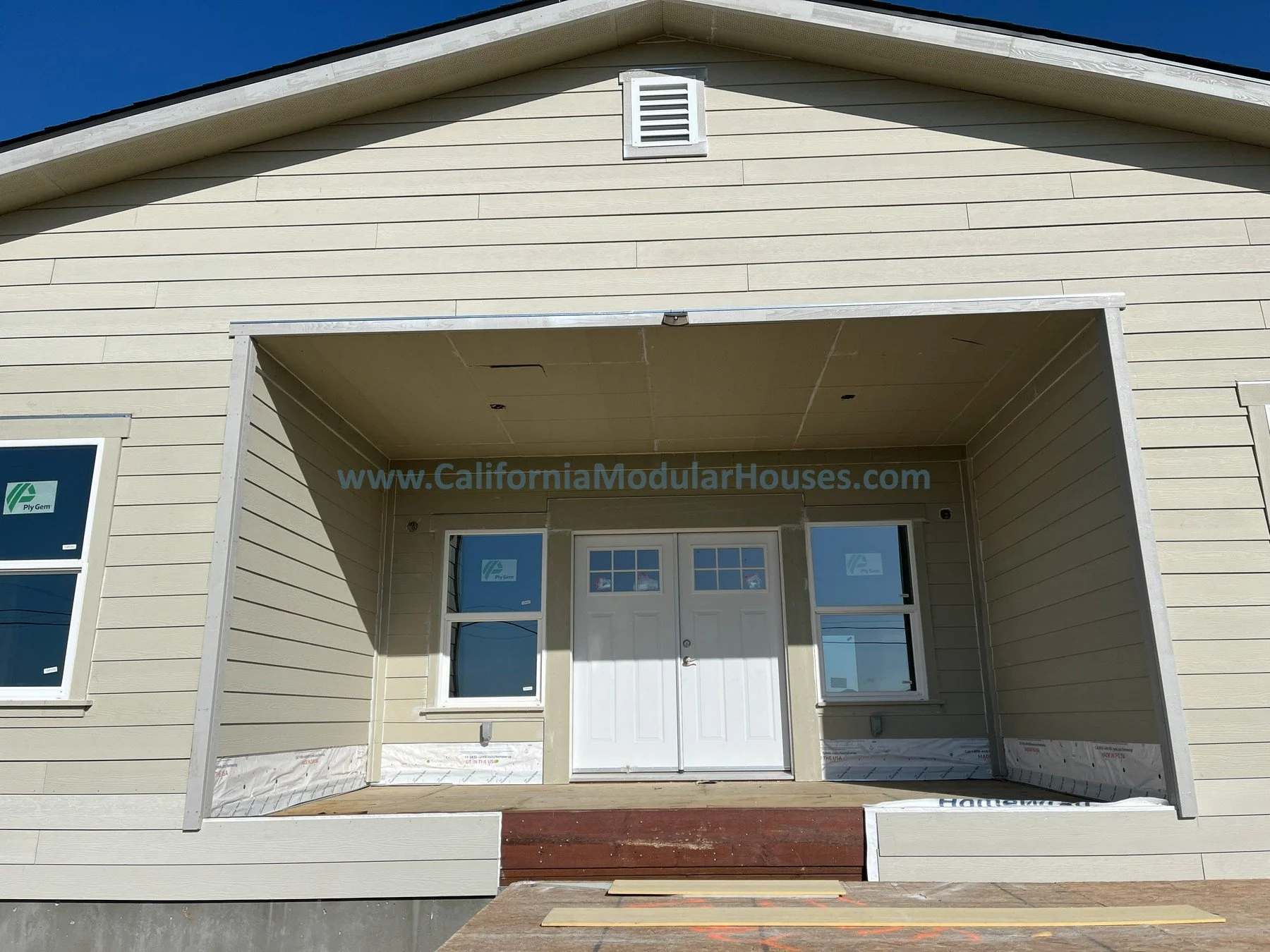 Front of a house under construction with a porch, beige exterior siding, white door with small windows, and two windows on each side of the door Exterior image L of a factory built home that has 3 bedrooms and 2 bathrooms California prefab modular CA