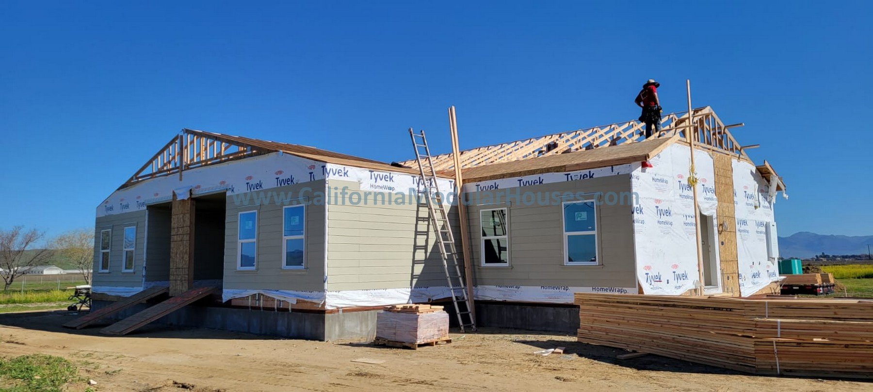 A house under construction with the roof framing being built, surrounded by stacks of wood and a ladder leaning against it, under a clear blue sky.  Roof of this modular home being completed on-site by general building contractor. 