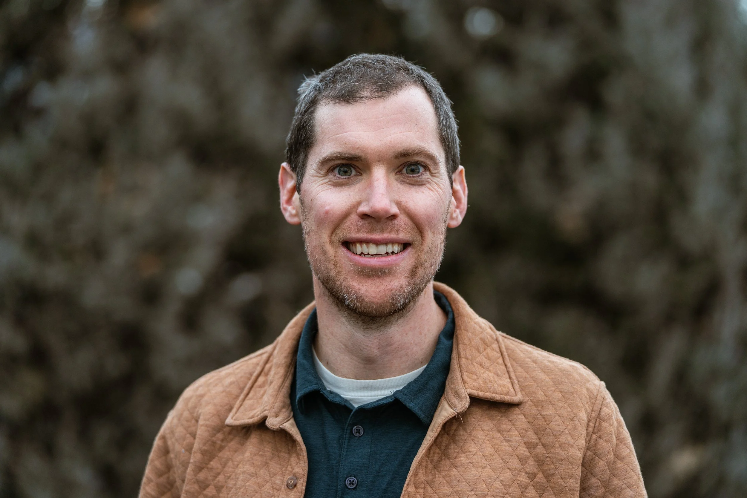 A smiling man with short brown hair and light skin stands outdoors in front of a blurred natural background, wearing a brown quilted jacket over a dark collared shirt.