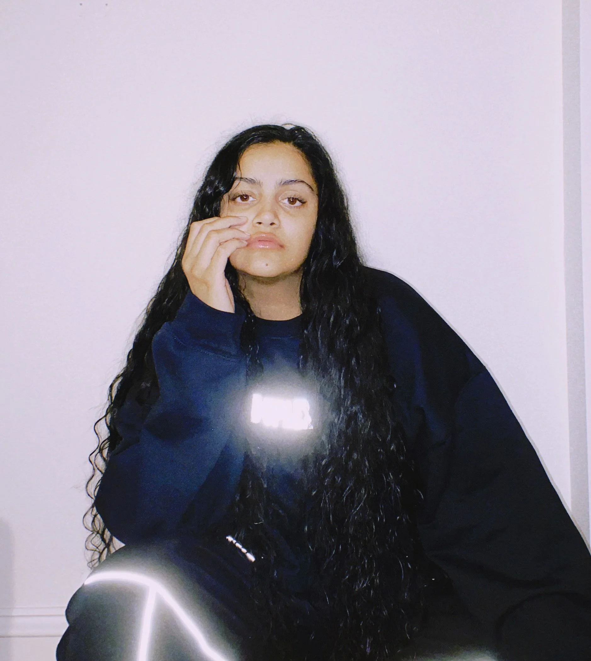 A young woman with long, curly black hair wearing a dark sweatshirt, sitting against a plain light-colored wall, looking at the camera with her hand resting on her face.
