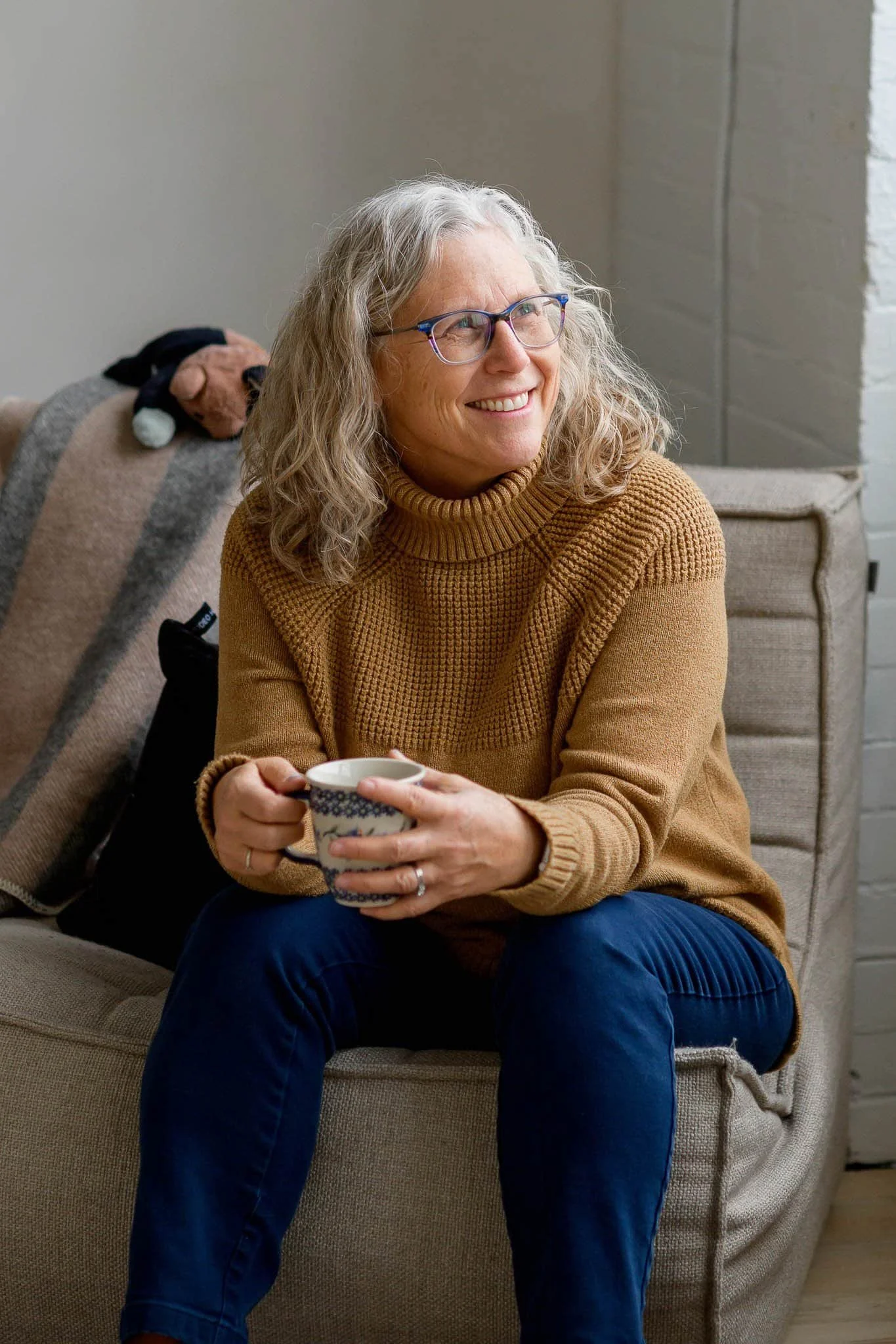 A woman with curly gray hair and glasses, smiling and holding a mug, sitting on a beige couch with a stuffed dog on the backrest.
