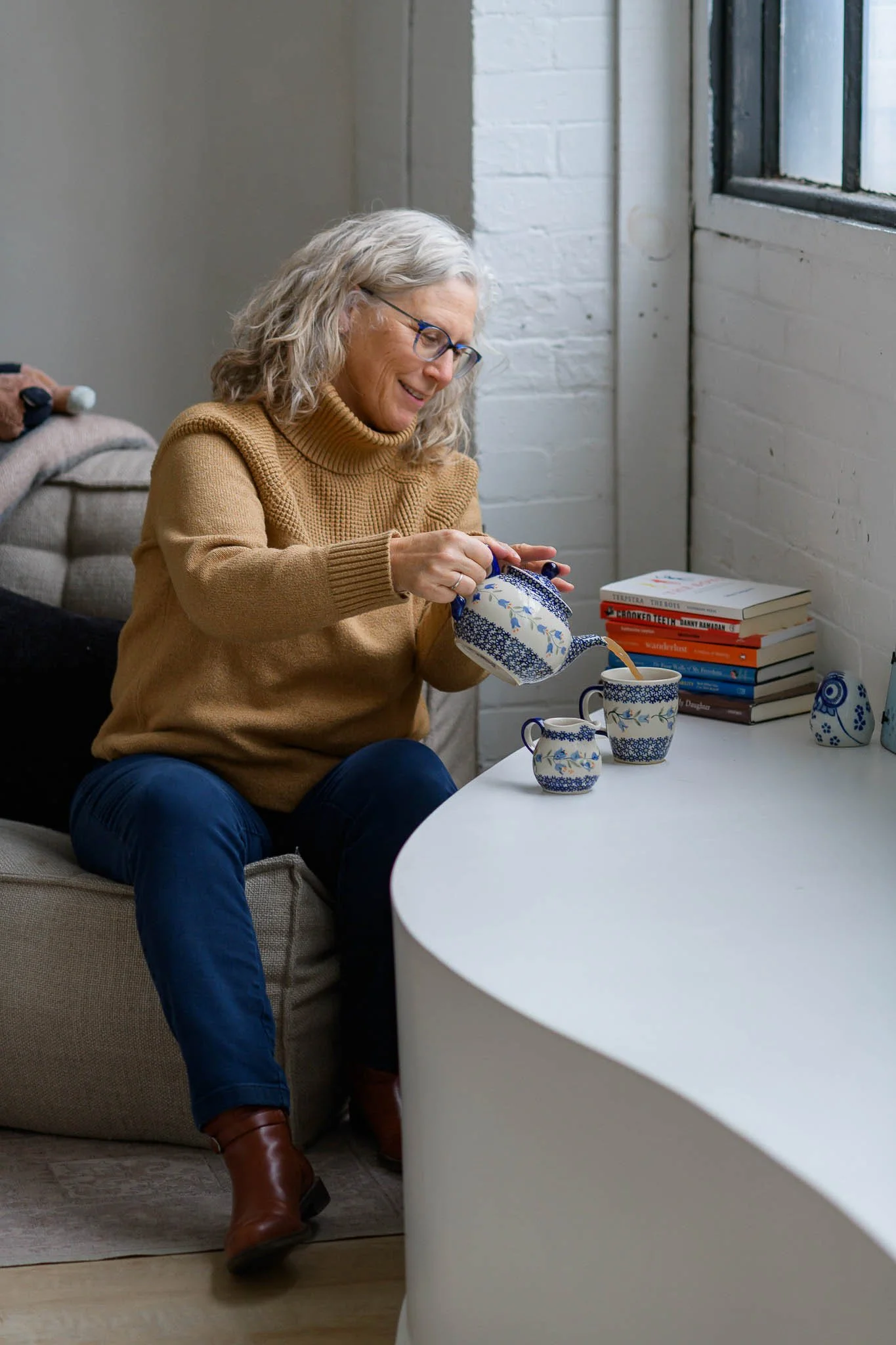 An elderly woman with gray hair and glasses, wearing a brown sweater and dark jeans, is sitting on a beige sofa, pouring tea from a patterned teapot into two matching cups at a white table, with a stack of books and small decorative objects nearby.