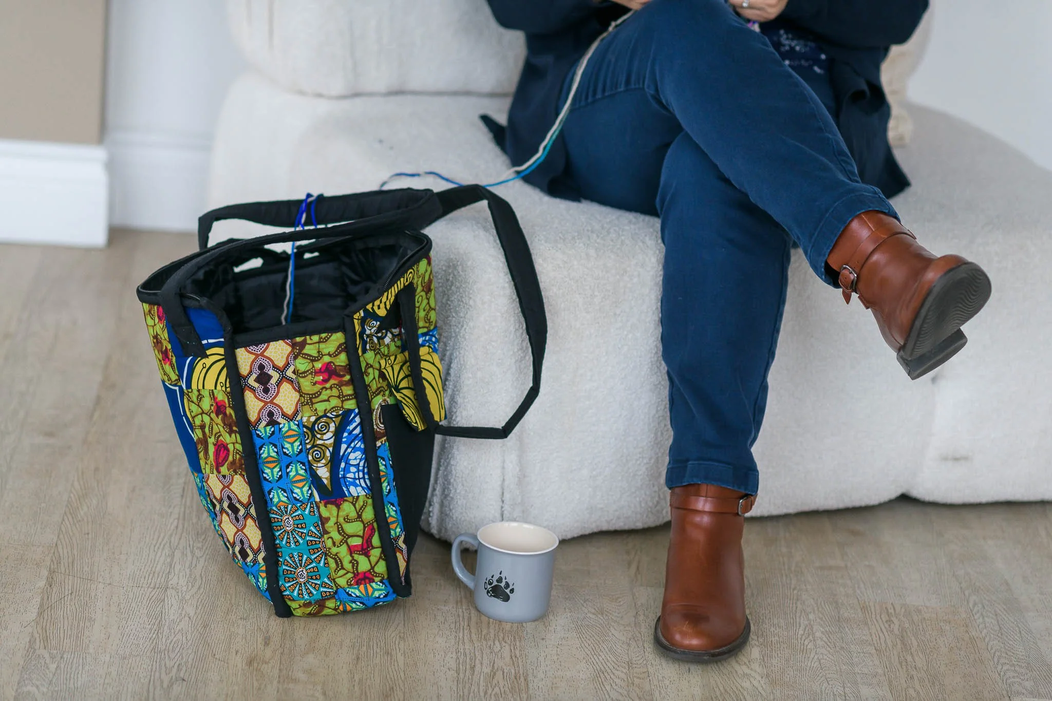 Person sitting on a light gray sofa with one leg crossed over the other, wearing blue jeans and brown boots. A colourful, patterned bag and a gray mug with a paw print are nearby on the floor.