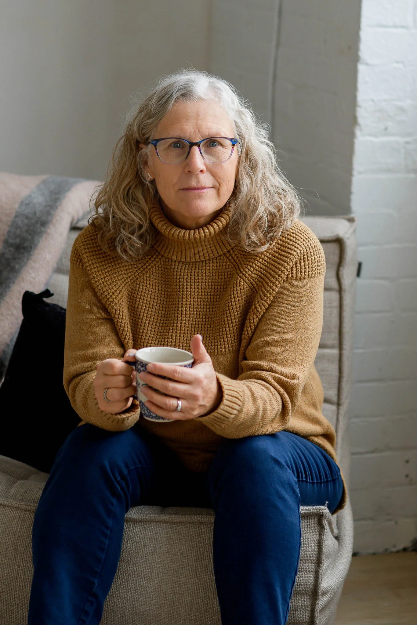A woman with curly gray hair and glasses sitting on a beige couch, holding a patterned mug, wearing a brown turtleneck sweater and blue pants, with a neutral indoor background.