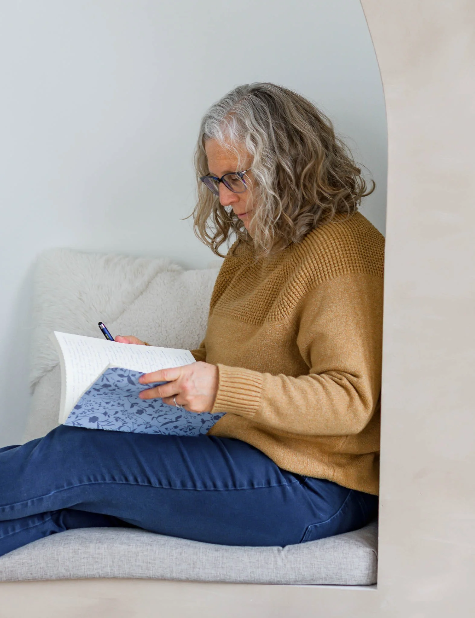 A woman with gray curly hair and glasses, wearing a brown sweater and blue pants, sitting on a beige cushion and reading a book with a blue floral cover in a cozy indoor setting.