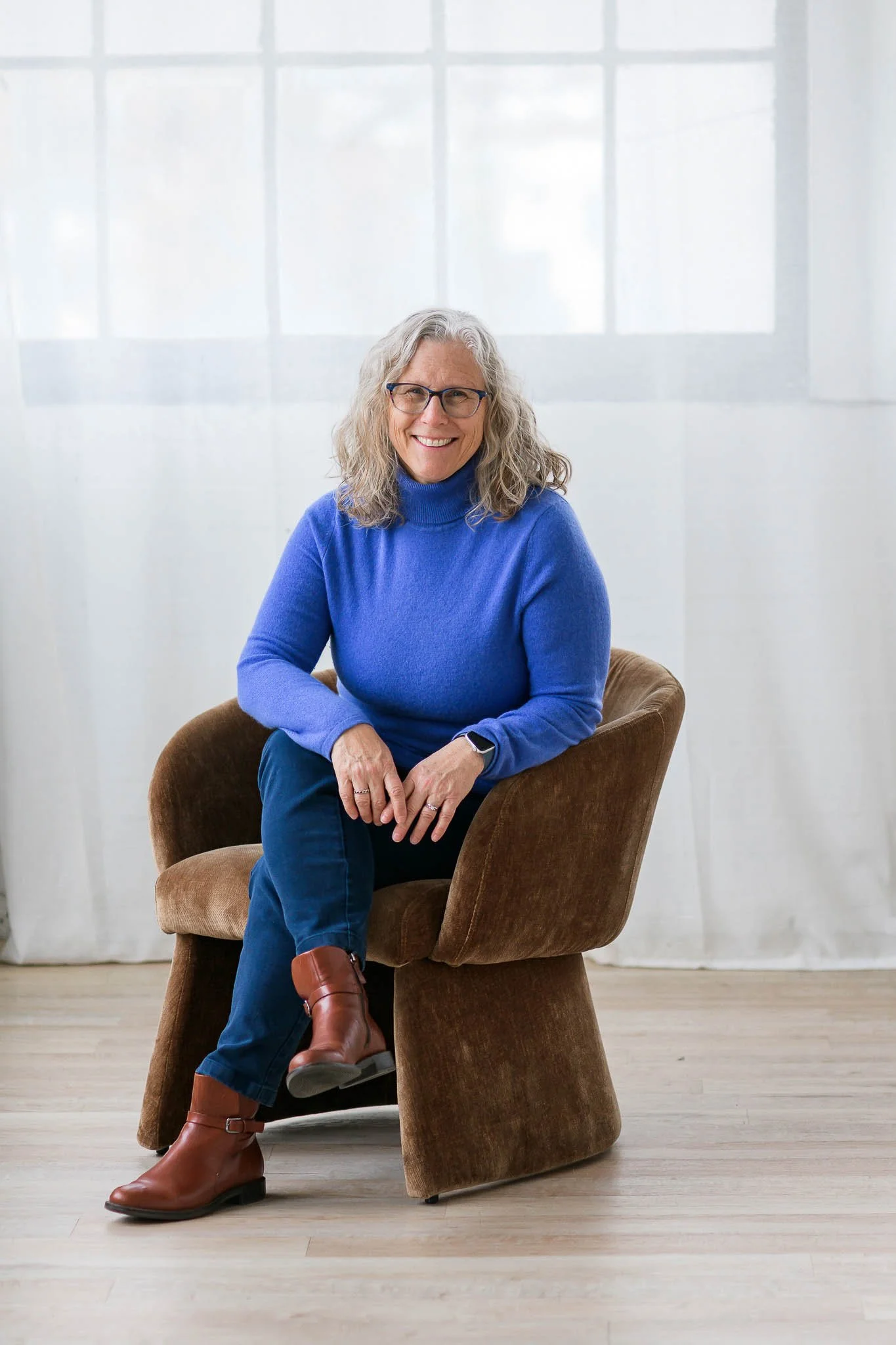 A woman with gray curly hair and glasses, wearing a blue turtleneck sweater and brown boots, sitting on a brown chair in front of a white curtain and window.