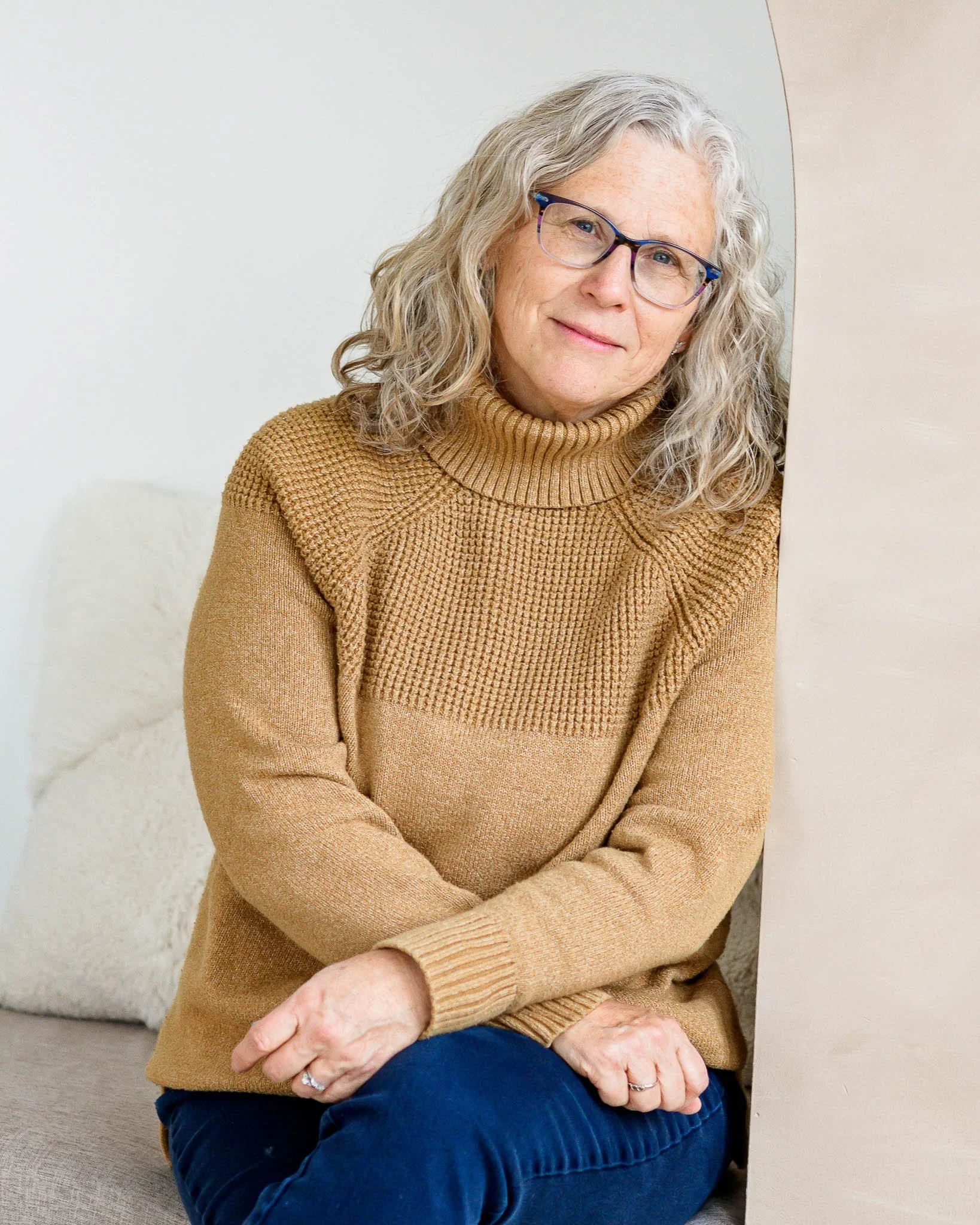 A woman with gray curly hair and glasses sitting on a beige couch, wearing a tan sweater and blue jeans, in a cozy indoor setting.