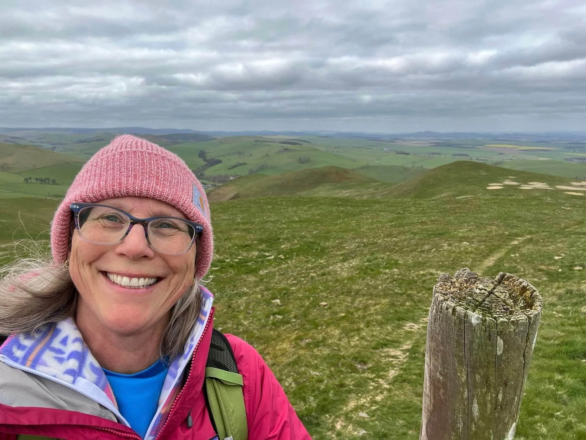 Smiling woman with glasses in pink hat and outdoor jacket taking a selfie on a grassy hill with rolling green hills and overcast sky in the background.